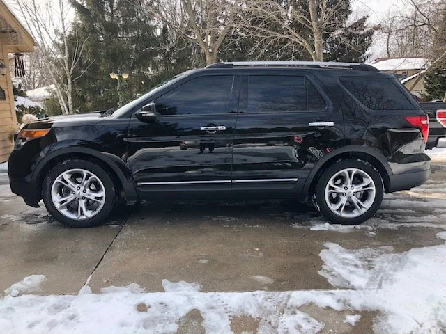 A black SUV parked on a driveway with snow and ice, with trees and houses in the background.