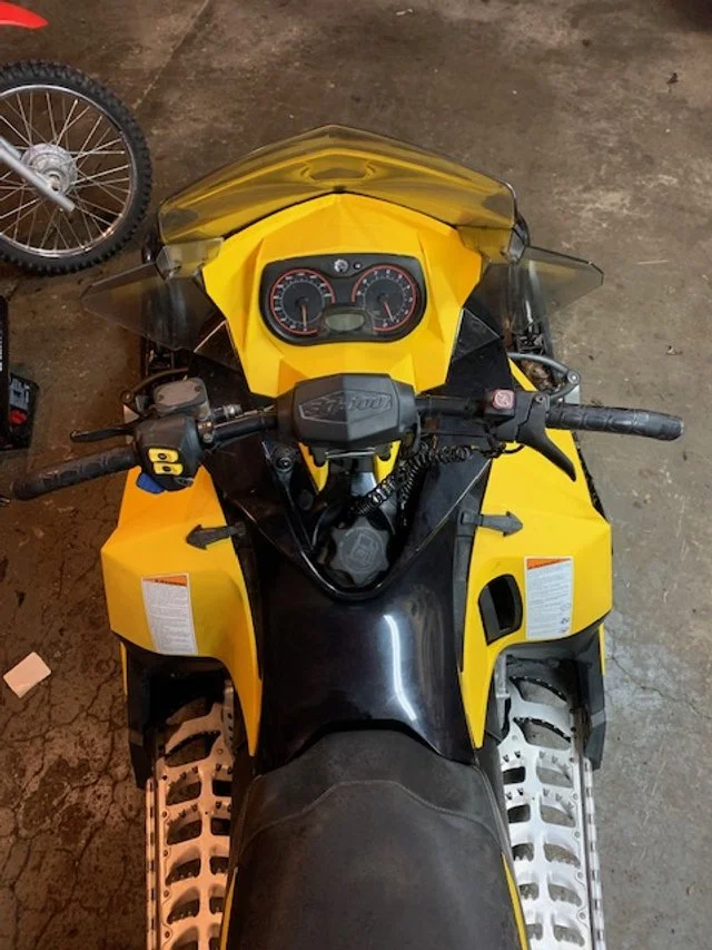 A yellow snowmobile with a black seat and dashboard, viewed from above, on a concrete floor.