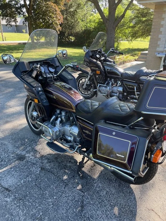Two black touring motorcycles parked on a driveway with a grassy lawn and trees in the background.