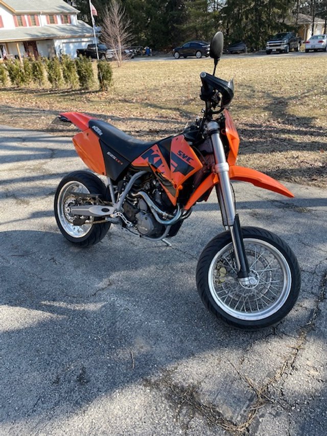 Orange KTM dirt bike parked on a paved surface with trees and cars in the background.