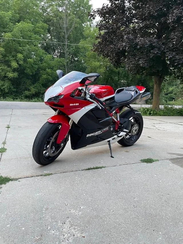 A red, black, and white Ducati superbike parked on a driveway with trees and greenery in the background.