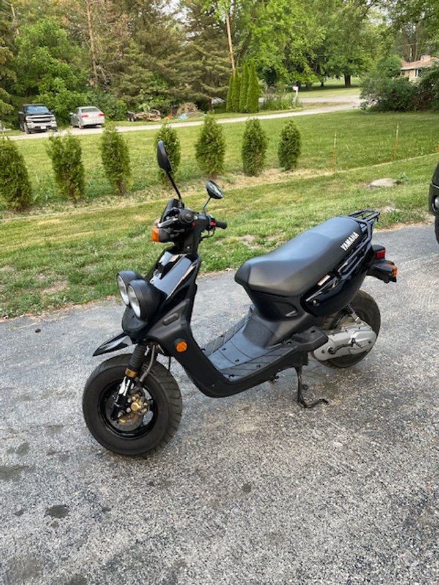 Black Yamaha scooter parked on a gravel driveway with grass and trees in the background.