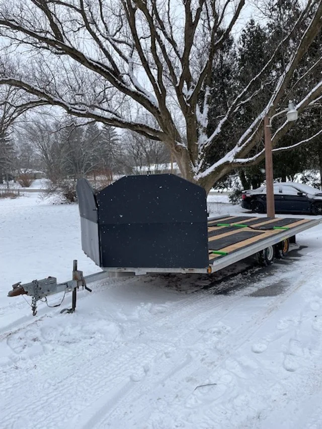 A flatbed trailer with a black enclosure parked on snow-covered ground near a large tree and a parked car in the background.