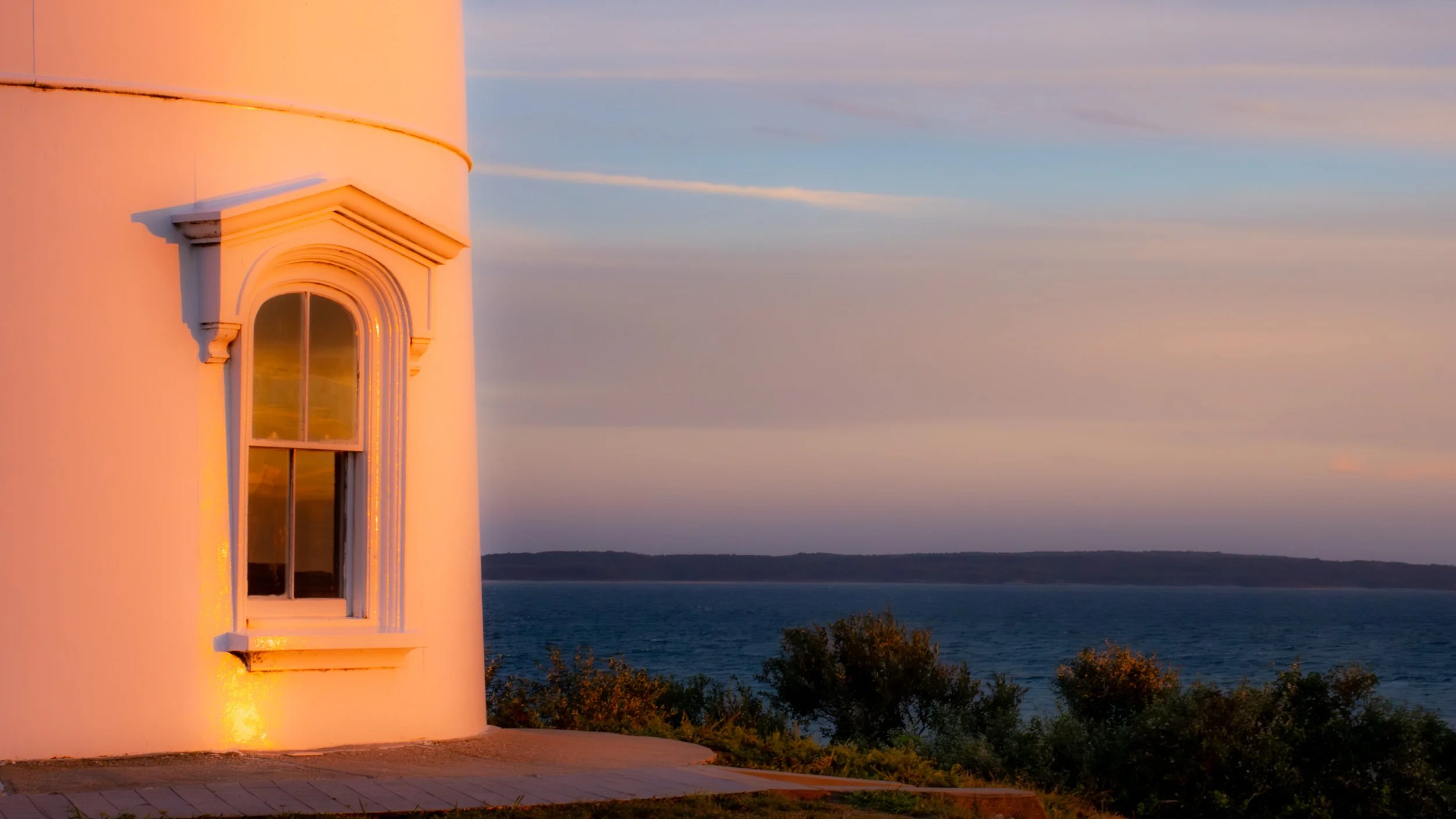 White lighthouse tower illuminated by warm light near sunset, with a window visible and the ocean in the background.