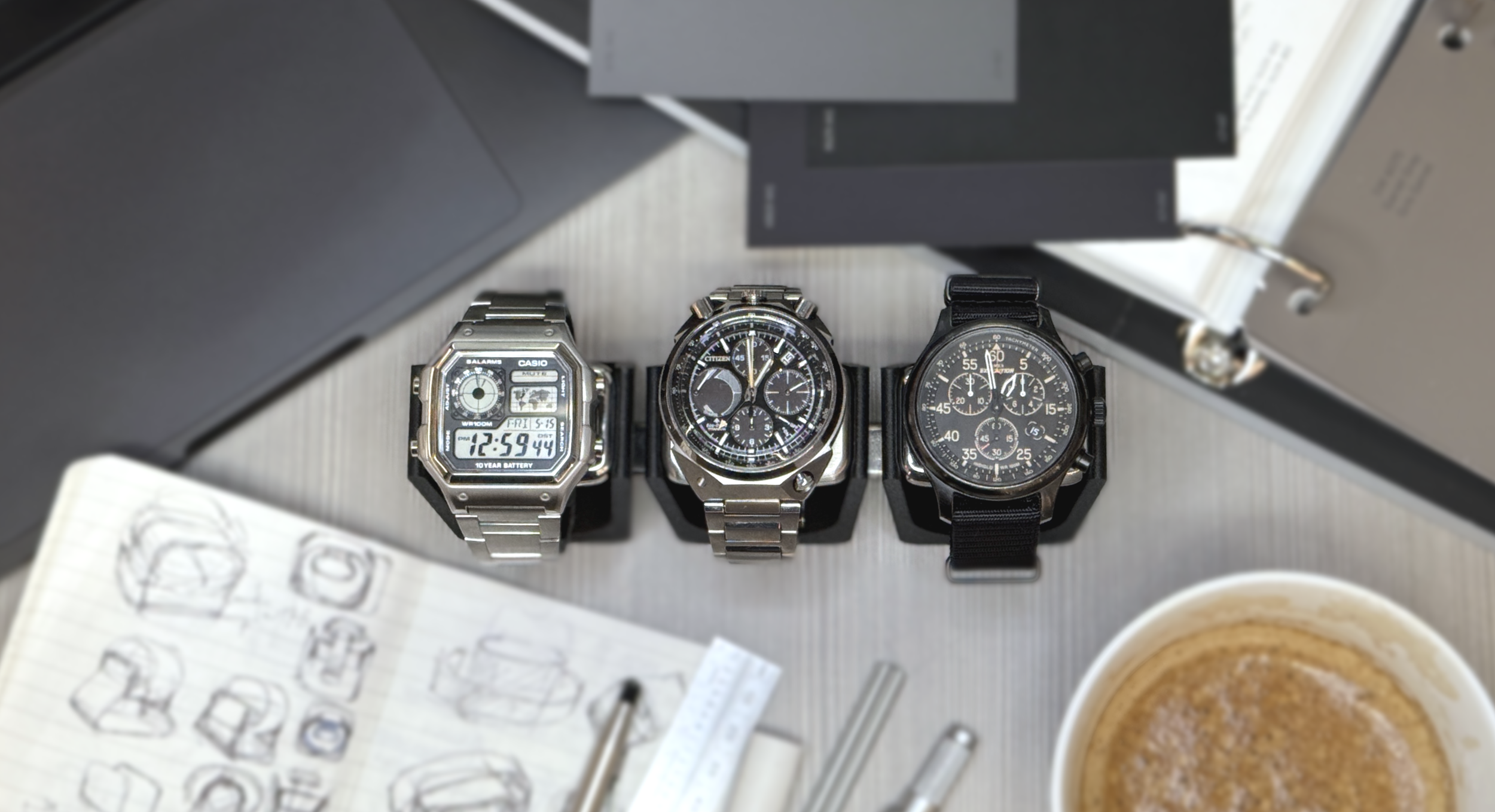 Three wristwatches with metal and leather bands arranged on a surface, with a notebook, sketches of watch designs, and a coffee cup nearby.