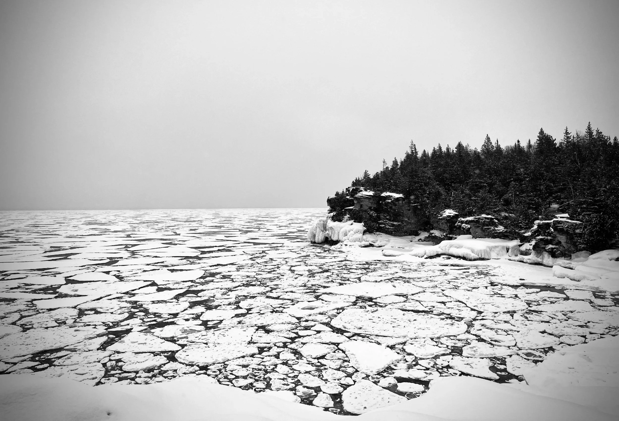 A black and white photograph of the frozen Bruce Peninsula shoreline. The iced lake is beginning to crack, revealing a distant forest to the right. The landscape serves as a metaphor for product development: a harsh, relentless winter that, through p