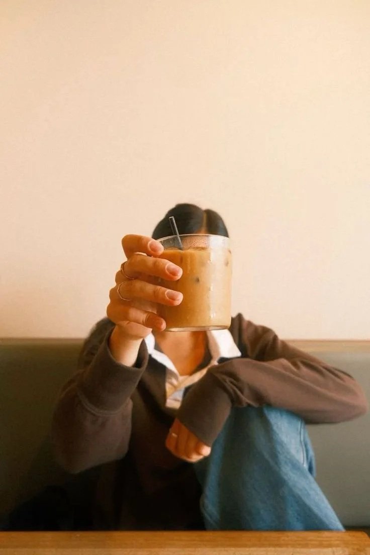 A person sitting at a table holding a glass of iced coffee or iced tea with a straw, in a cafe or restaurant setting.