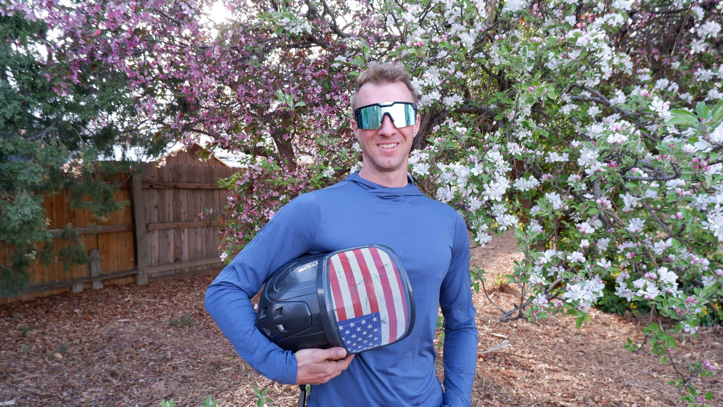 A smiling man in blue athletic clothing and reflective sunglasses holding a motorcycle helmet with a patriotic design of red and white stripes and a blue section with white stars, standing outdoors near a flowering tree with pink and white blossoms.