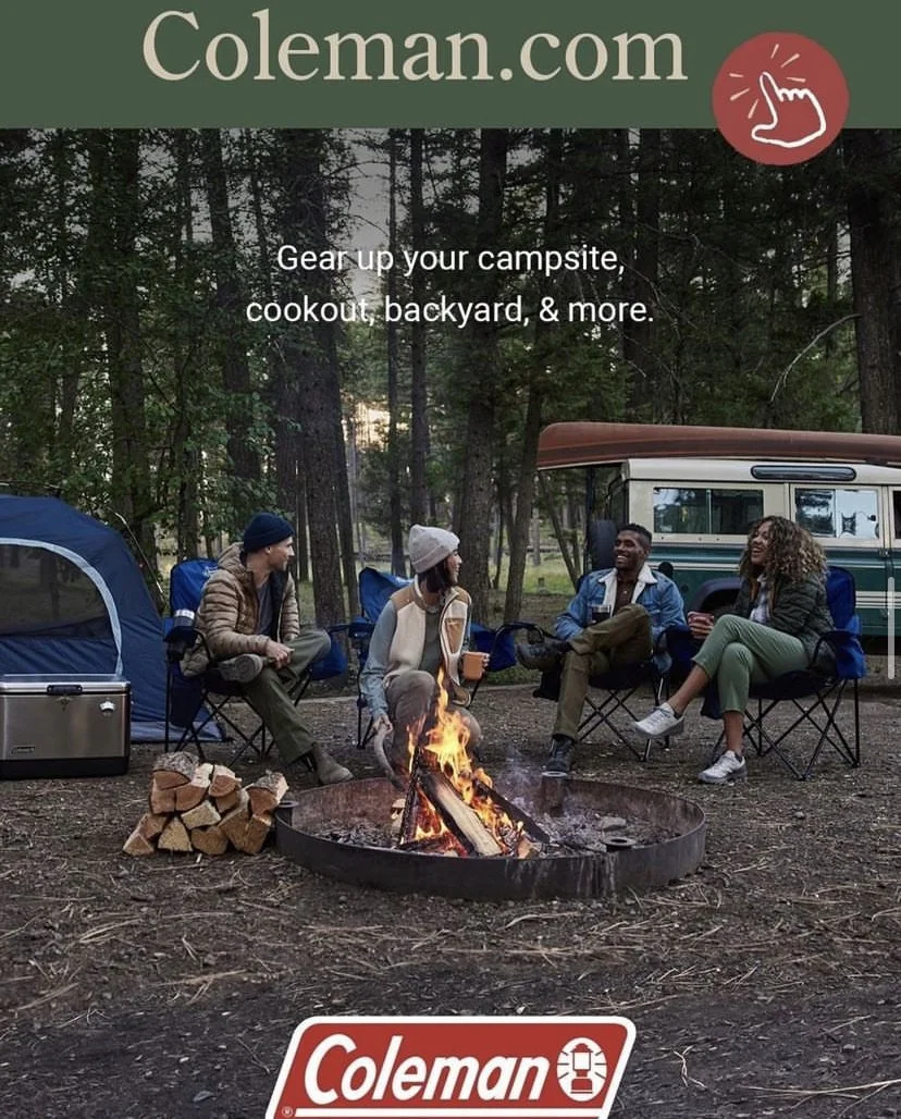 Four people sitting around a campfire in a forest, with camping equipment and a camper van nearby.