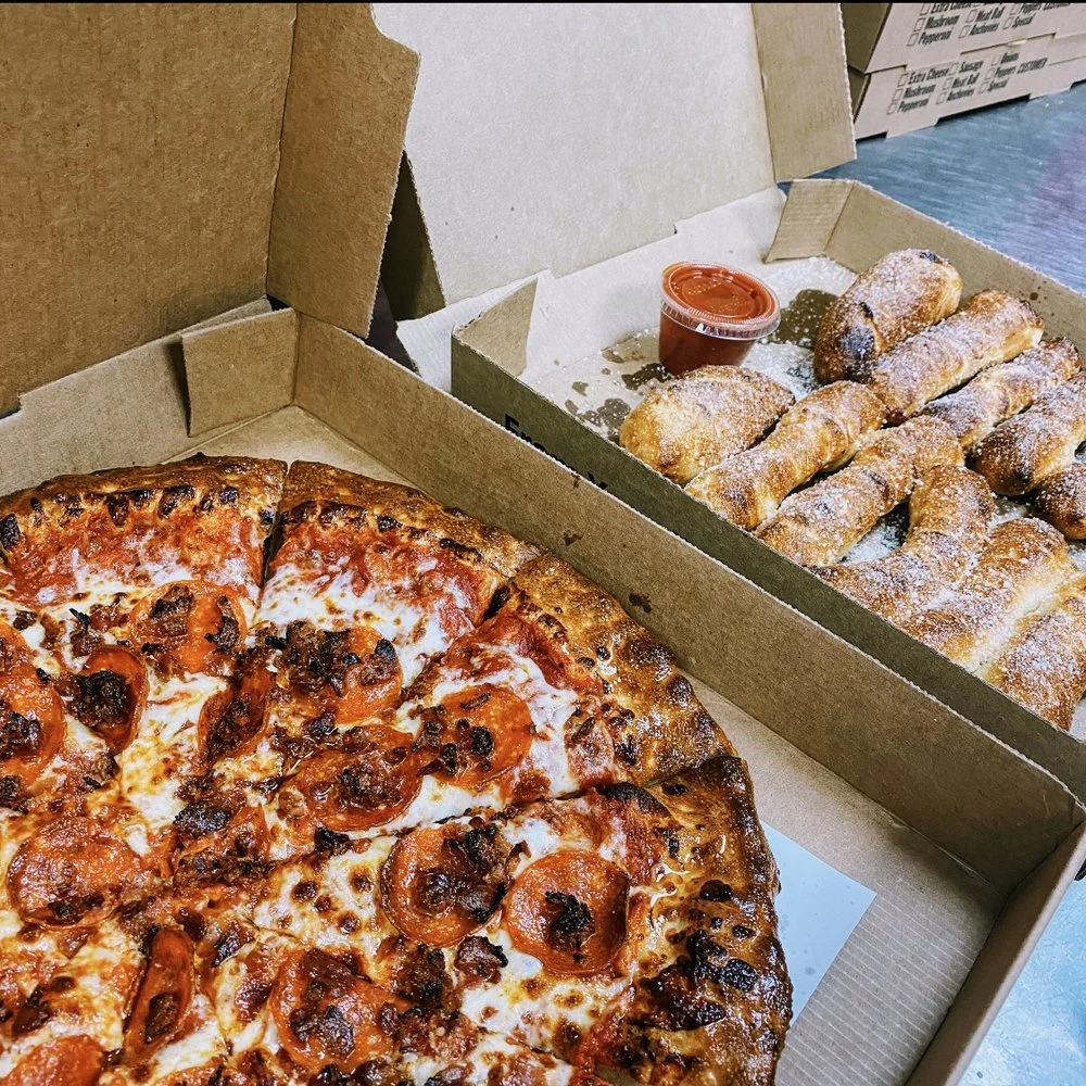 A pizza with pepperoni and sausage in a pizza box next to a tray of powdered sugar-dusted cannoli with a small cup of red dipping sauce.