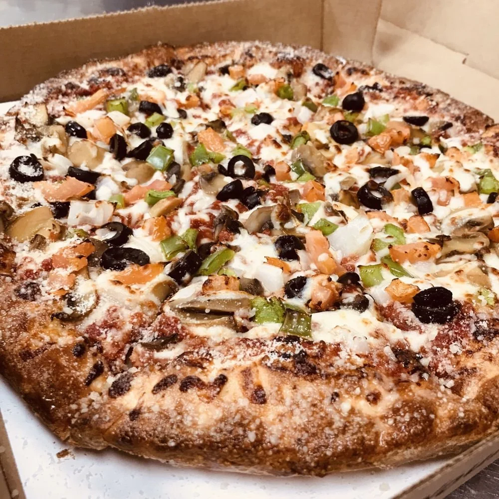 A rectangular pizza with a thick crust topped with cheese, black olives, green bell peppers, mushrooms, and diced tomatoes in a cardboard pizza box.