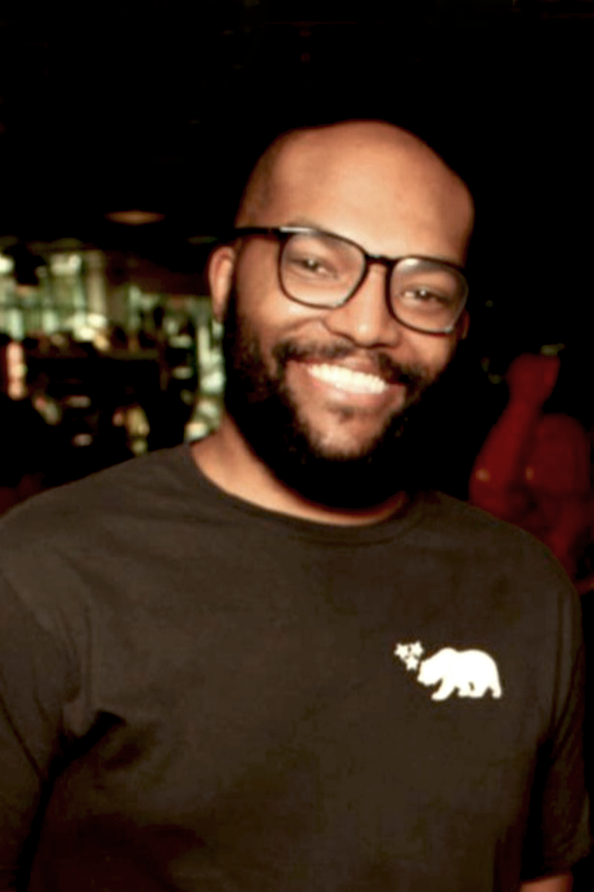 A smiling man with glasses, a beard, and a mustache wearing a black t-shirt with a California bear logo with a star on its back, in what appears to be a public indoor setting.