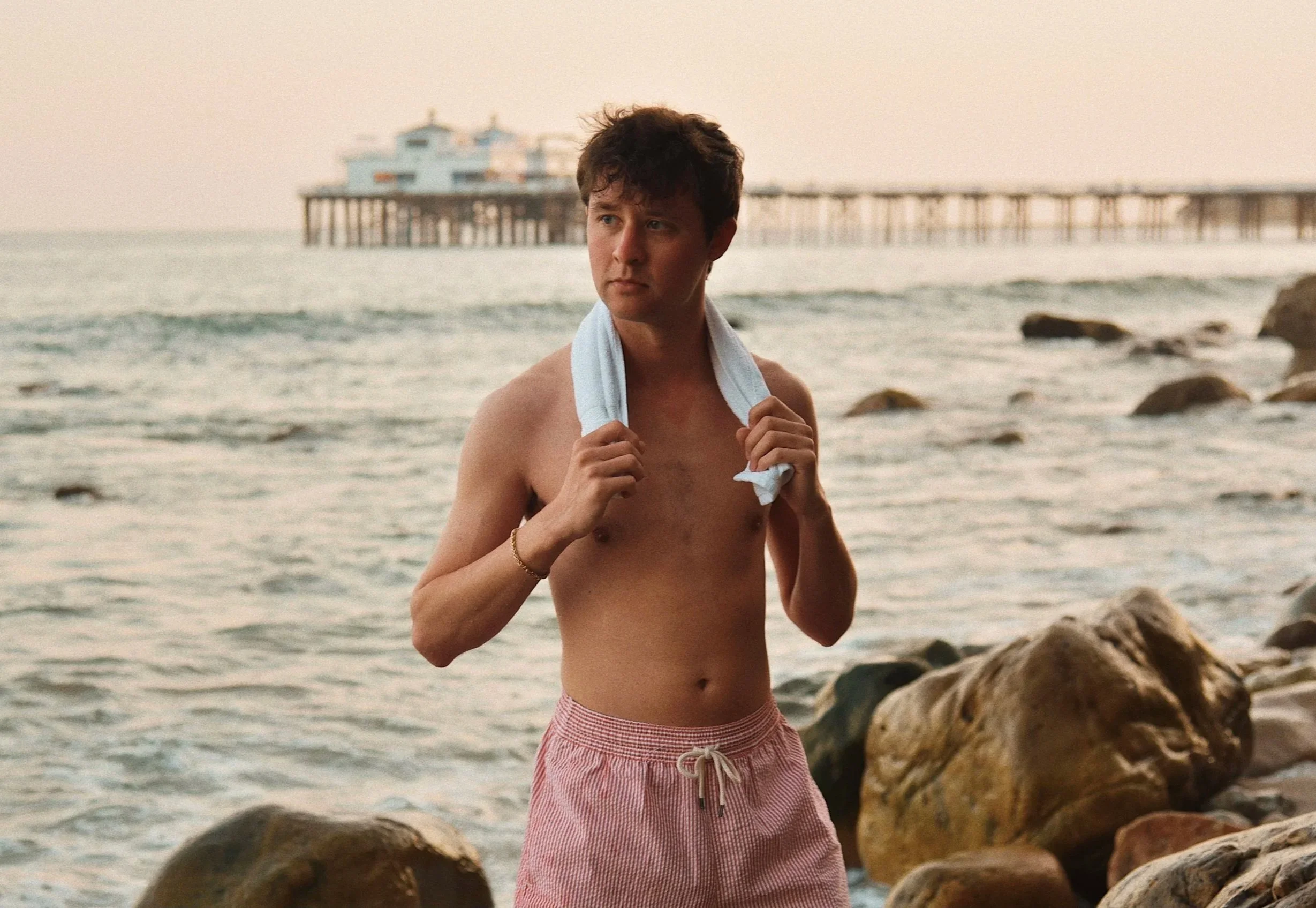 Kevin Severin in red shorts holding a towel around his neck, standing on a rocky beach with a pier and a building in the background during sunset.