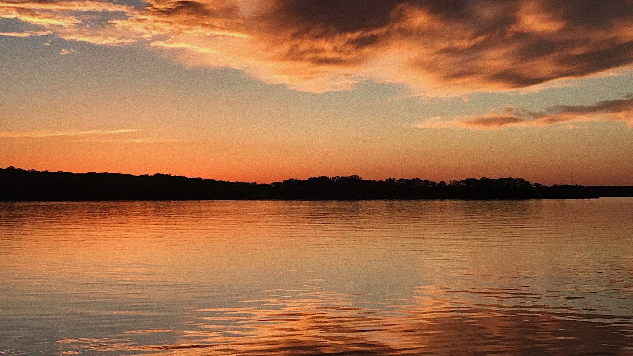 Reflections of a sunset on a body of water with a peninsula and low hanging clouds in the background.