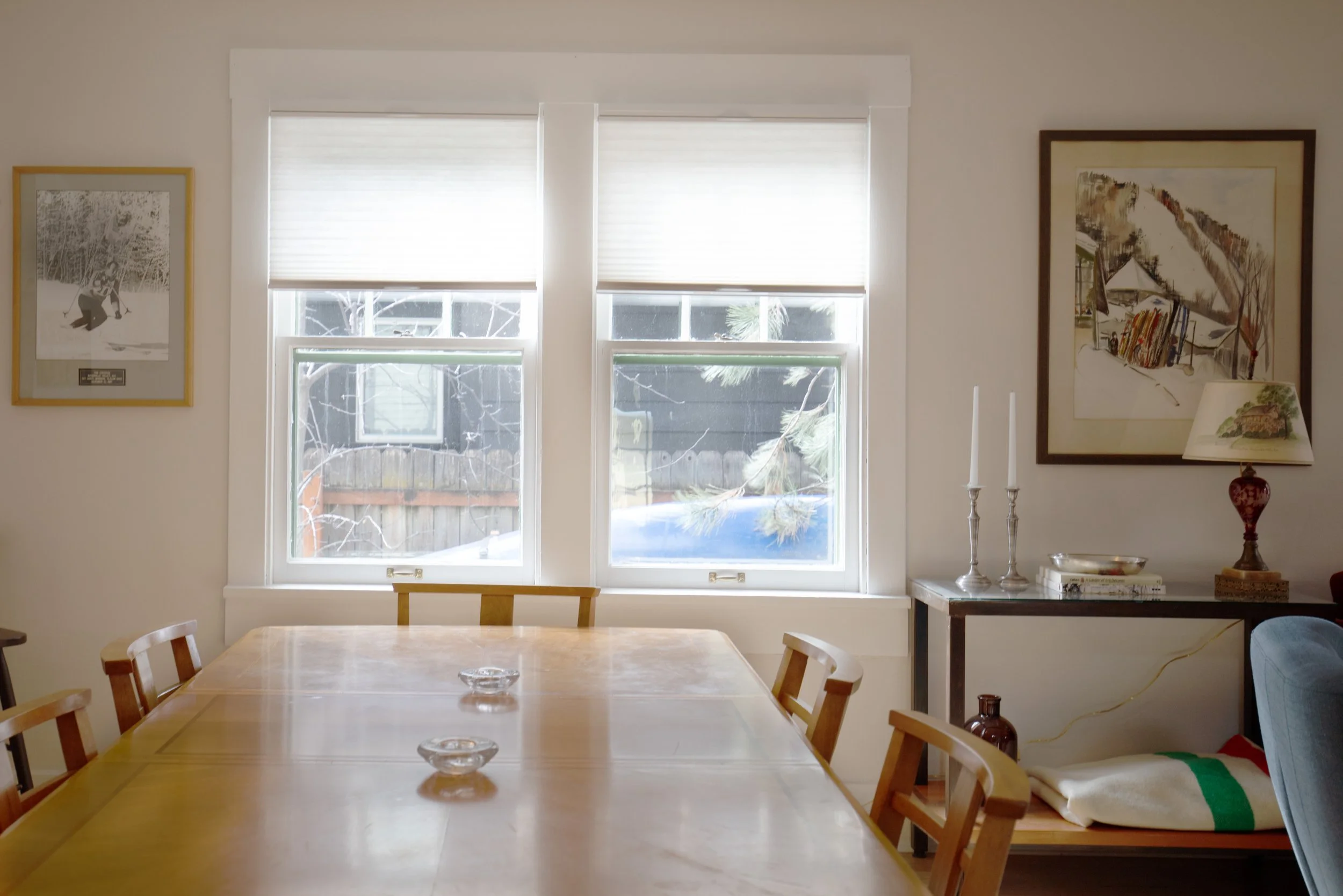 A dining room with a wooden table and six chairs, two bowls on the table, two framed pictures on the white walls, a side table candles, books, and a decorative lamp, and a window with blinds allowing natural light. two dot interiors