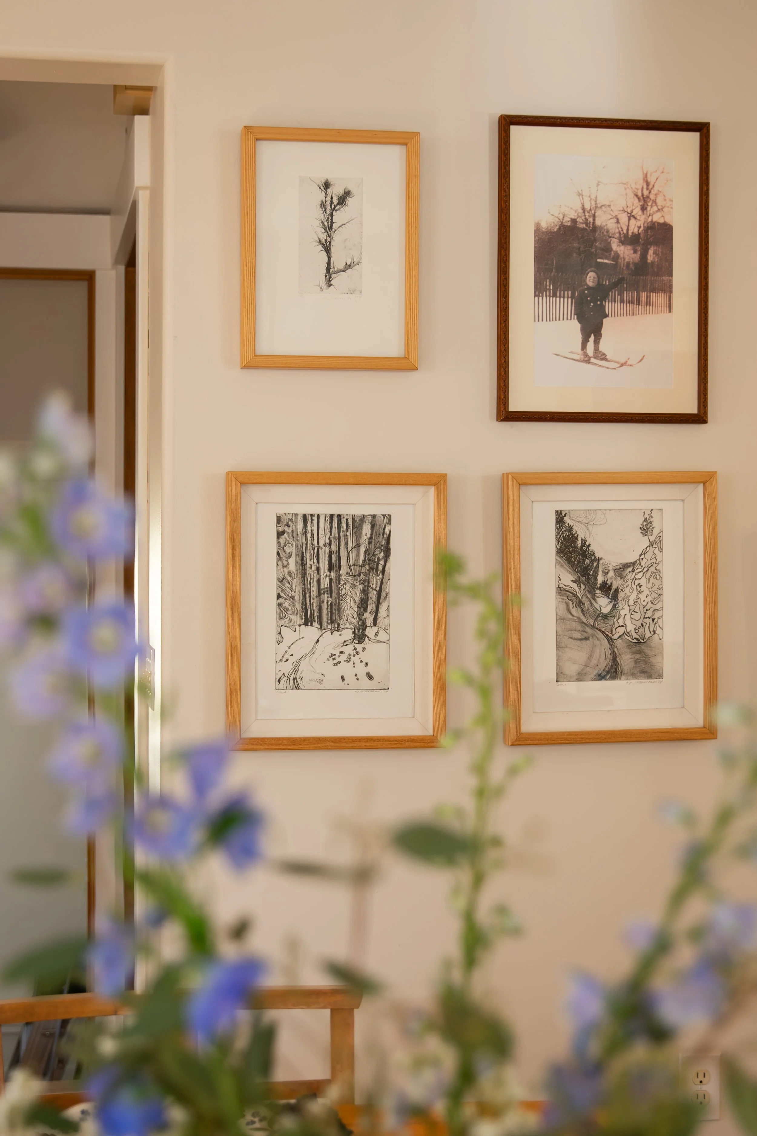 A wall decorated with five framed pictures, including black-and-white sketches and a photograph of a child skiing, with some purple flowers and green leaves blurred in the foreground.