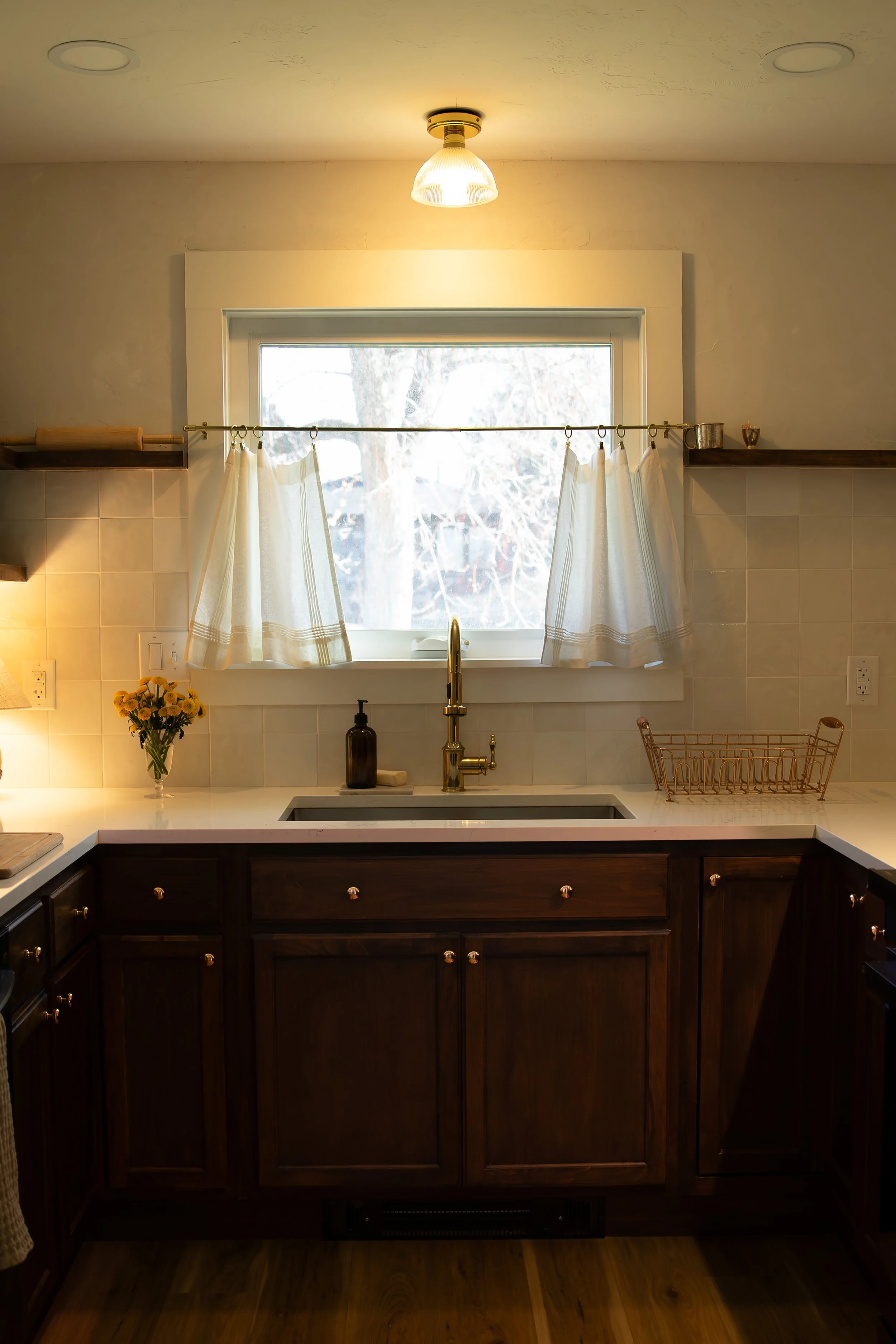 A cozy kitchen with a window above the sink, featuring a brass faucet, a soap dispenser, and a flower vase, illuminated by a ceiling light. Bozeman interior design, interior renovation, and interior styling. 