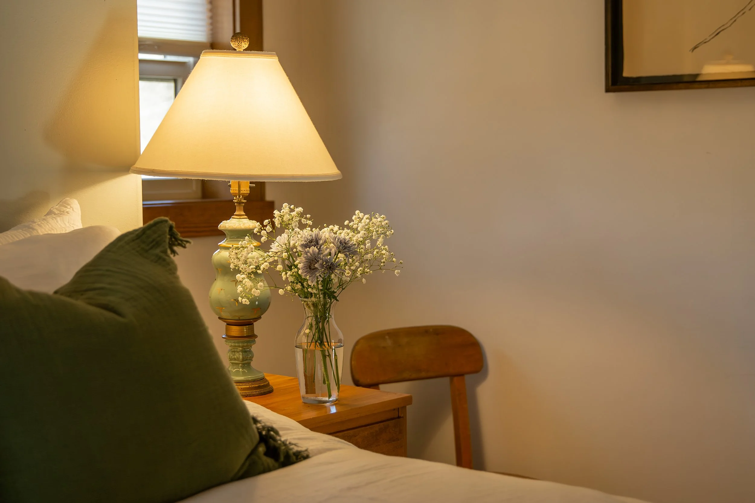 A bedroom corner with a wooden nightstand holding a lamp and a vase of flowers, a bed with pillows, and a wooden chair, with a window in the background.