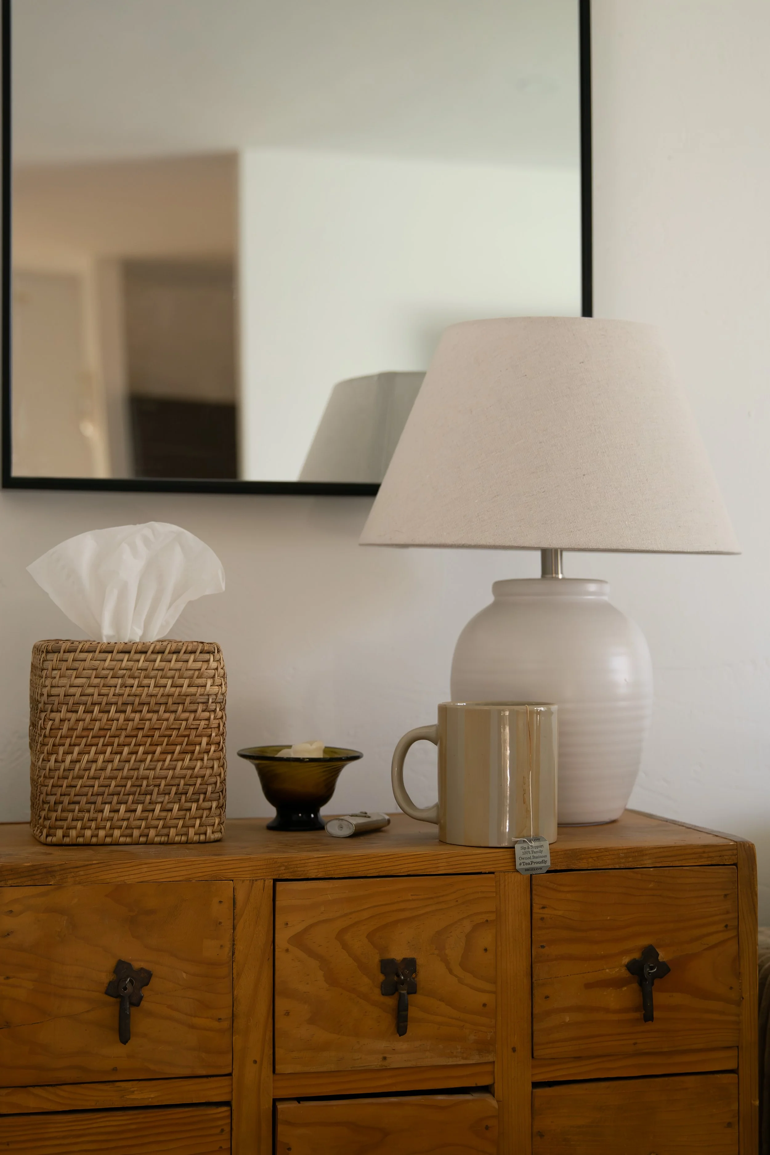 A wooden dresser with black metal handles topped with a white lamp, and a tissue box with tissues; a mirror above reflects part of an indoor space. Bozeman interior design, interior renovation, and interior styling. 