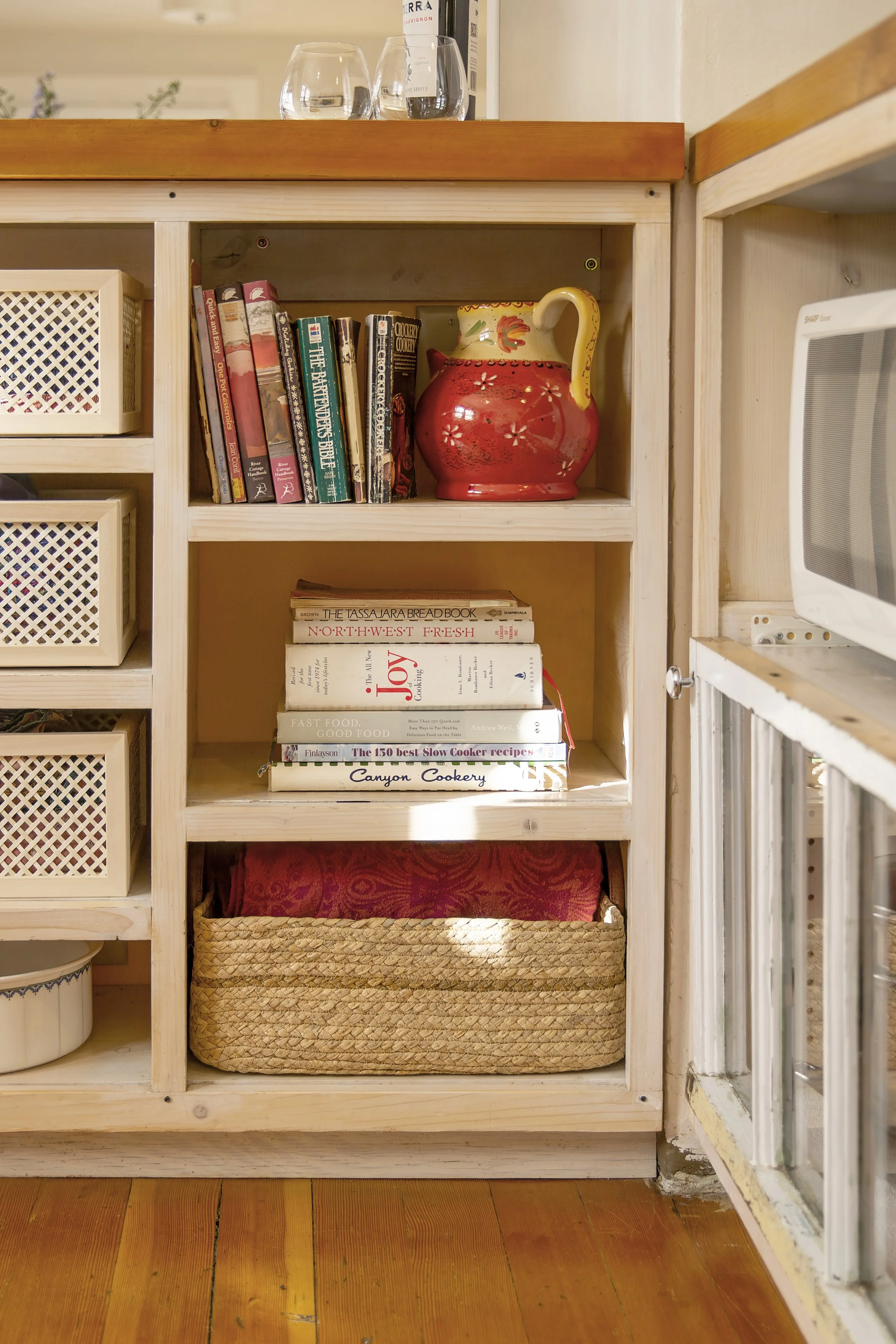 Wooden kitchen shelving with cookbooks, a decorative red and yellow ceramic pitcher, and baskets on a wooden floor. two dot interiors tidiness serves