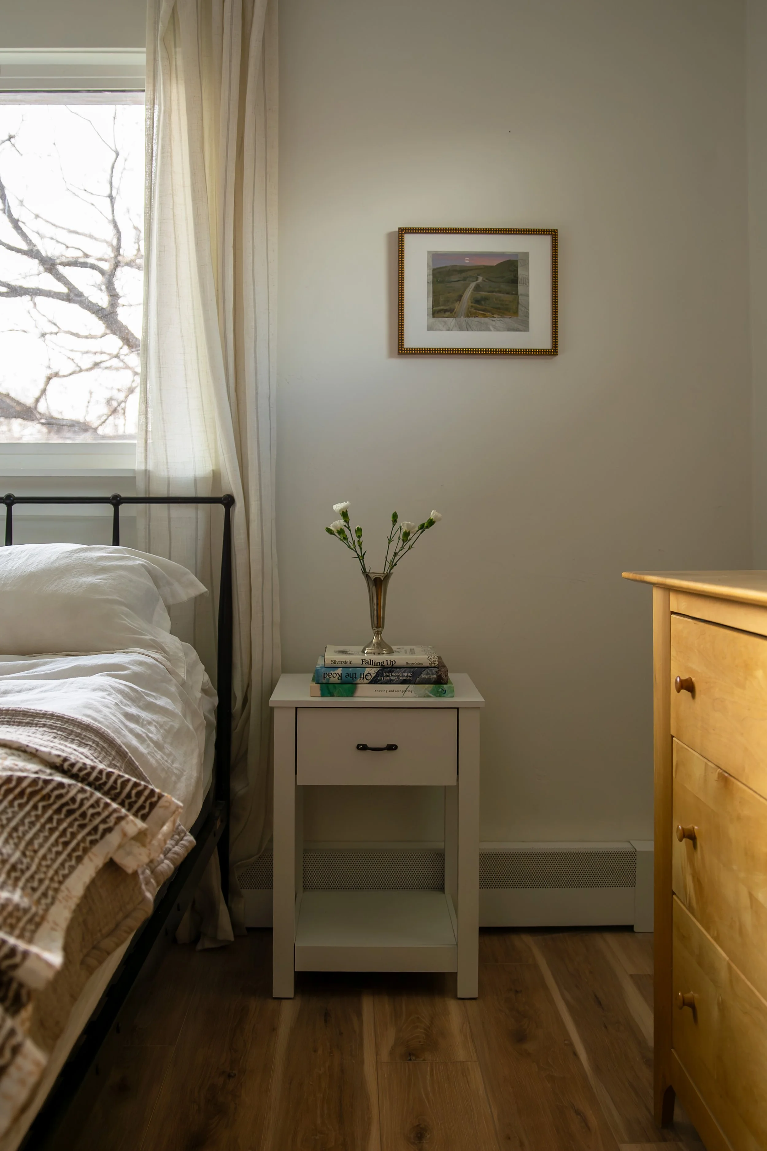 A bedroom with a white nightstand, a small vase with white flowers, books, a bed with white bedding, a window with curtains, and a framed picture on the wall. Bozeman interior design, interior renovation, and interior styling.