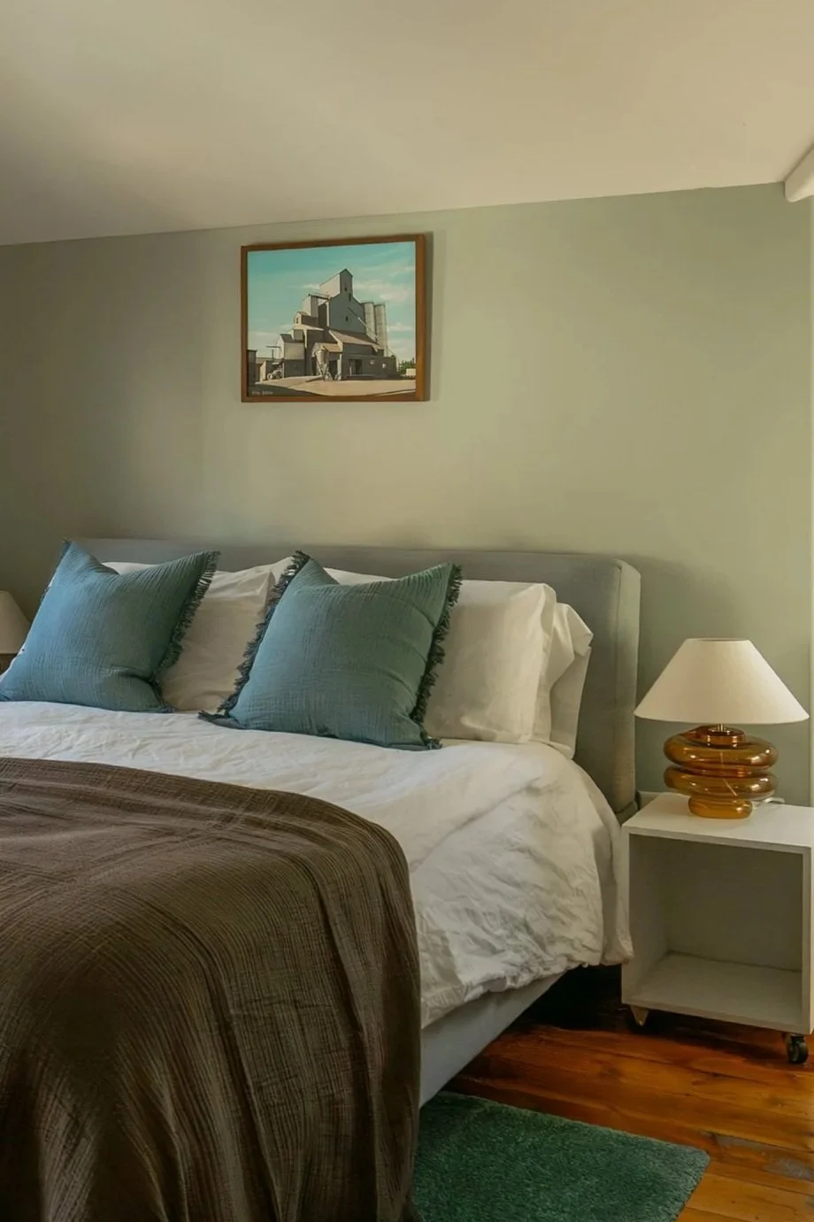 A neatly made bed with light sheets, three teal pillows with dark trim, a brown blanket, a white bedside table, a decorative amber-colored lamp, and a painting of a building on the wall in a cozy bedroom.   Bozeman Montana Interior Design Affordable