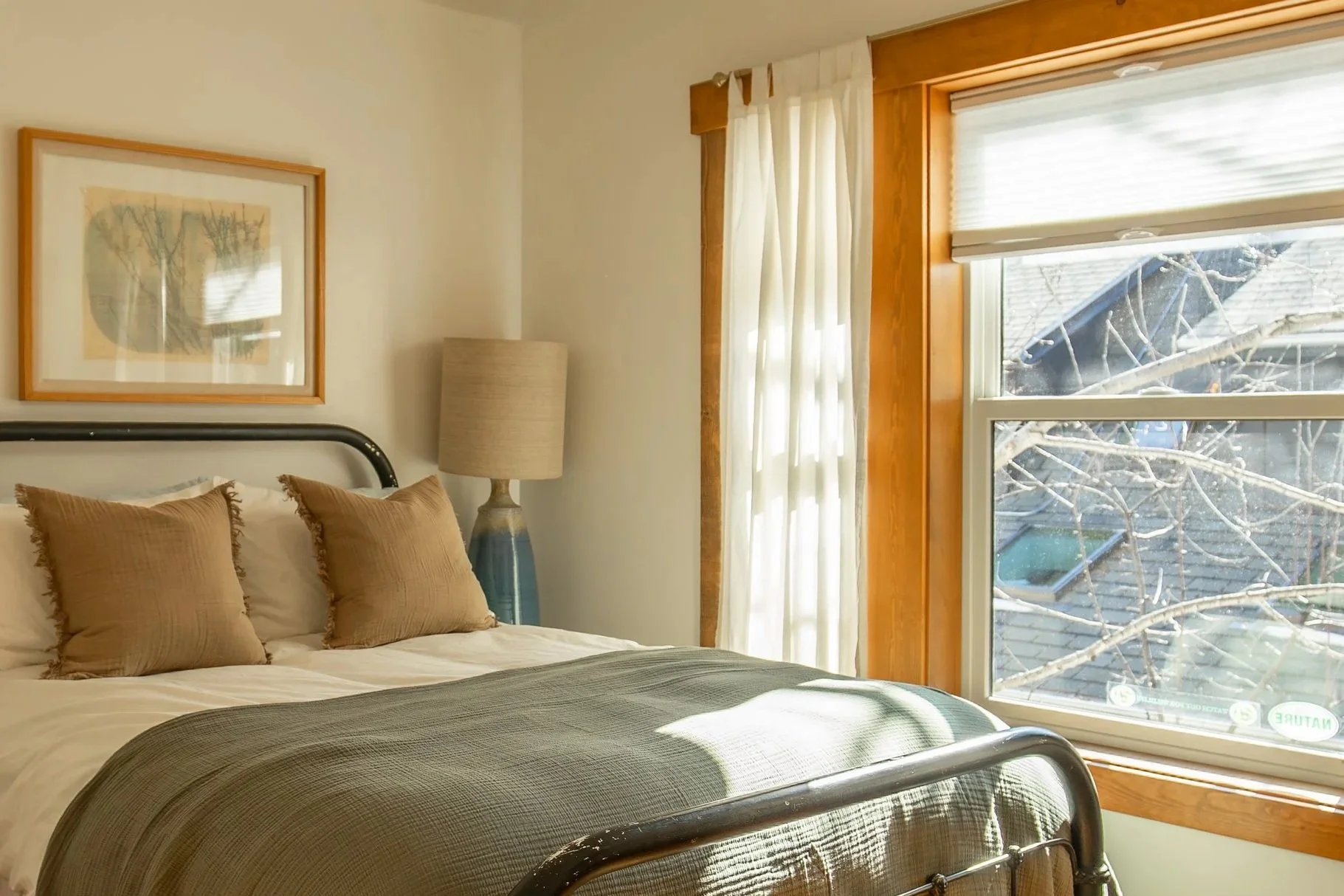A neatly made bed with three beige pillows, a green bedspread, in a bedroom with a window showing tree branches outside, a framed picture on the wall, and a tall lamp beside the bed.