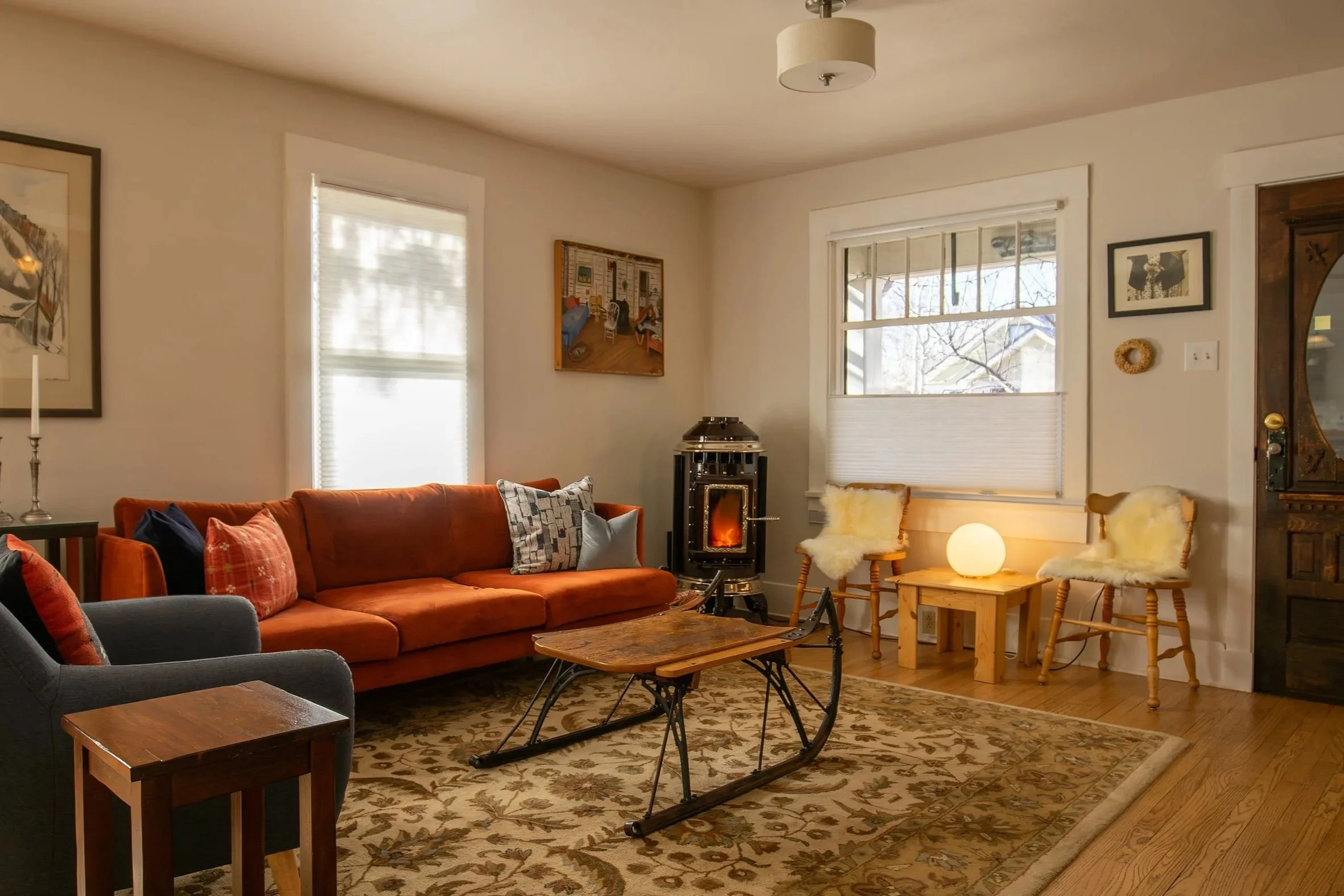 A cozy living room with an orange sofa, a black armchair with a plaid pillow, a wooden sled coffee table, a wood stove, two wooden chairs with fur cushions, a small wooden side table with a glowing round lamp, a patterned rug, and walls decorated wit