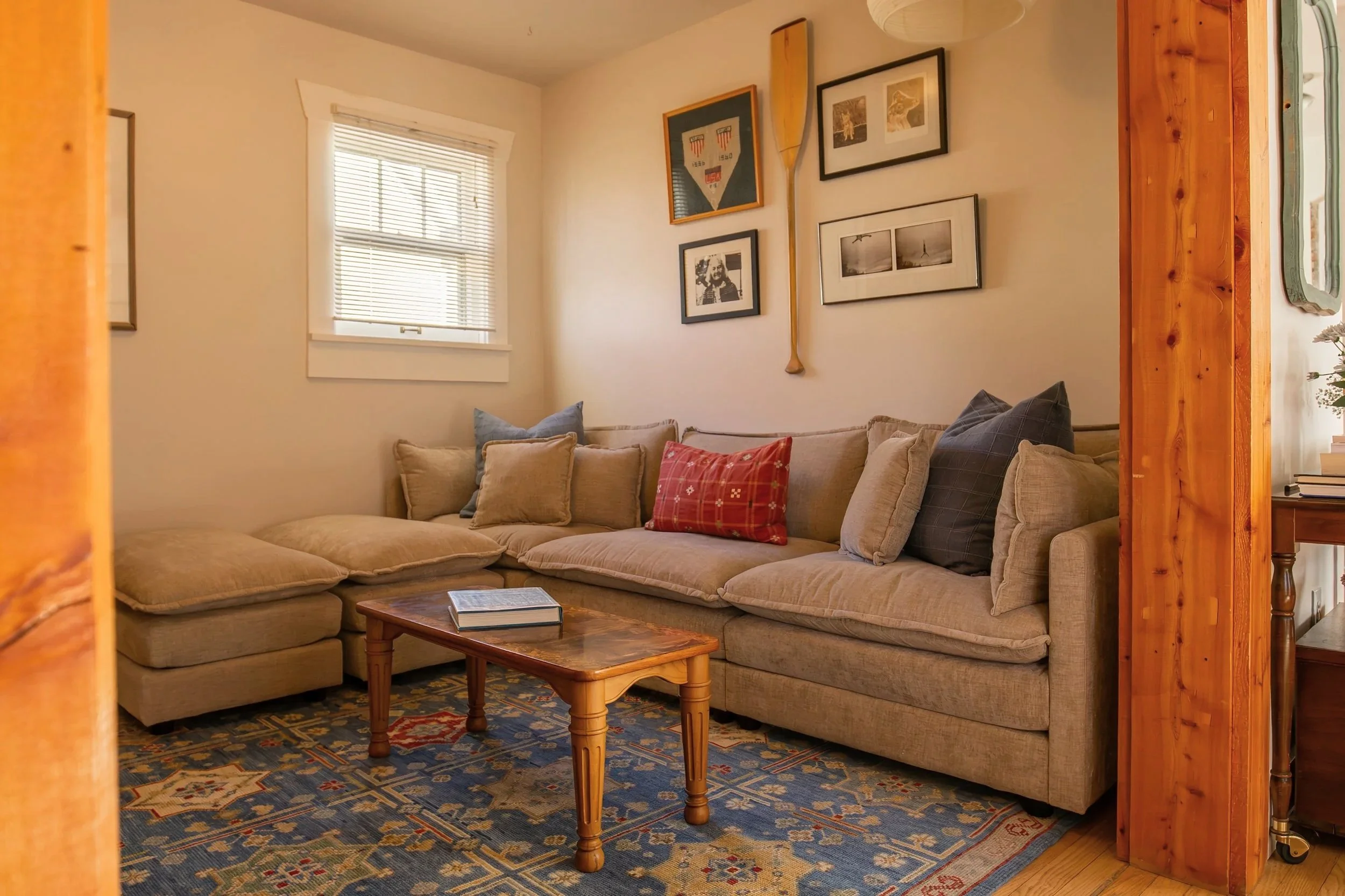 Living room with a beige sectional sofa, colorful cushions, a wooden coffee table with a book, a window with blinds, framed art on the wall, and a patterned rug. Bozeman Montana Affordable Interior Design gallery wall