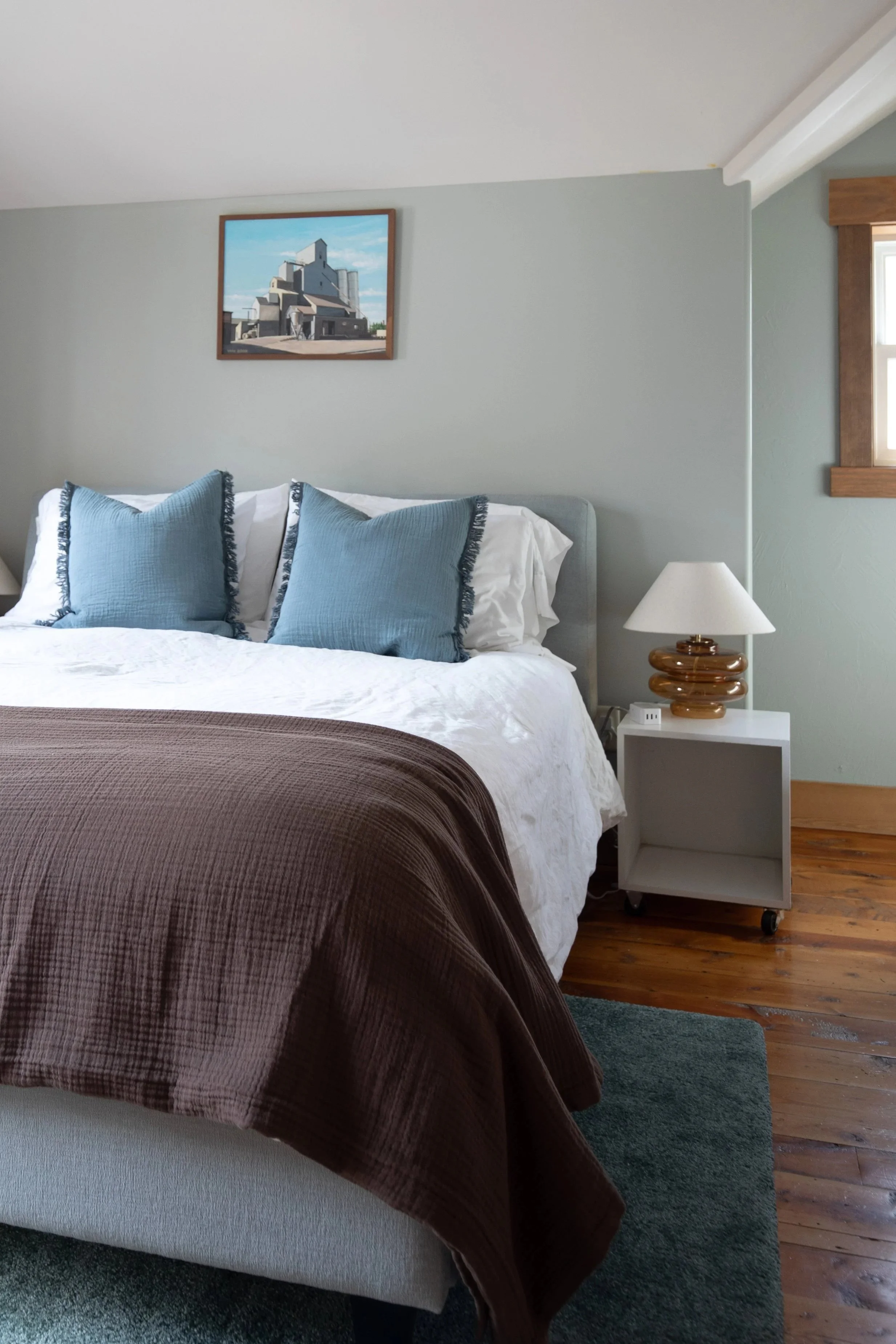 A cozy bedroom with a white bedspread and two large blue pillows. There is a small white nightstand with a vintage-style table lamp and a window with wooden trim on the right. A framed painting of a building hangs on the wall above the bed, and a dar