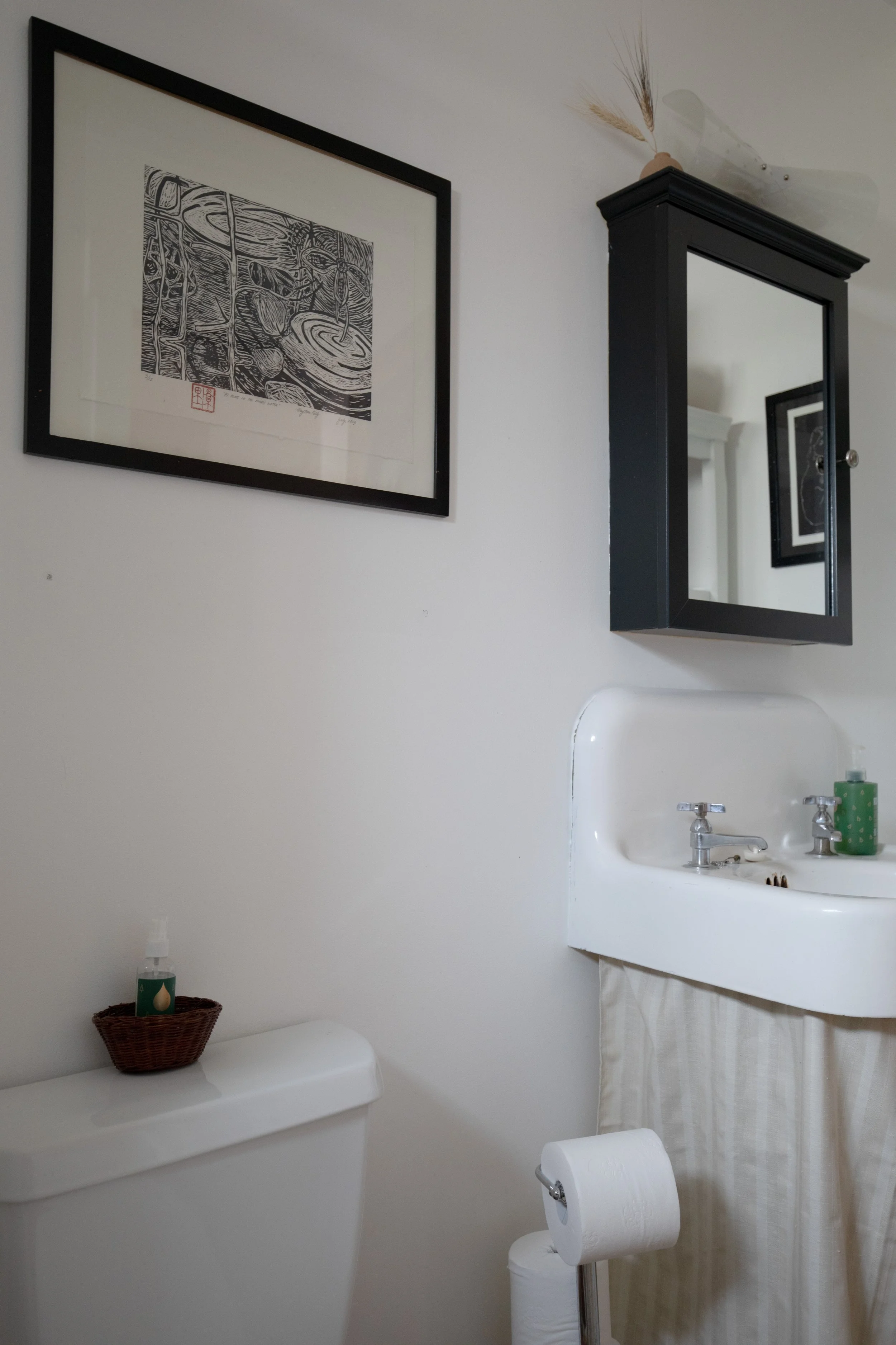 A small bathroom with a white toilet, a mini sink vintage, a black framed mirror, a framed black-and-white art print on the wall, and a roll of toilet paper on a holder. refresh renovation remodel interior designer bozeman montana two dot interiors