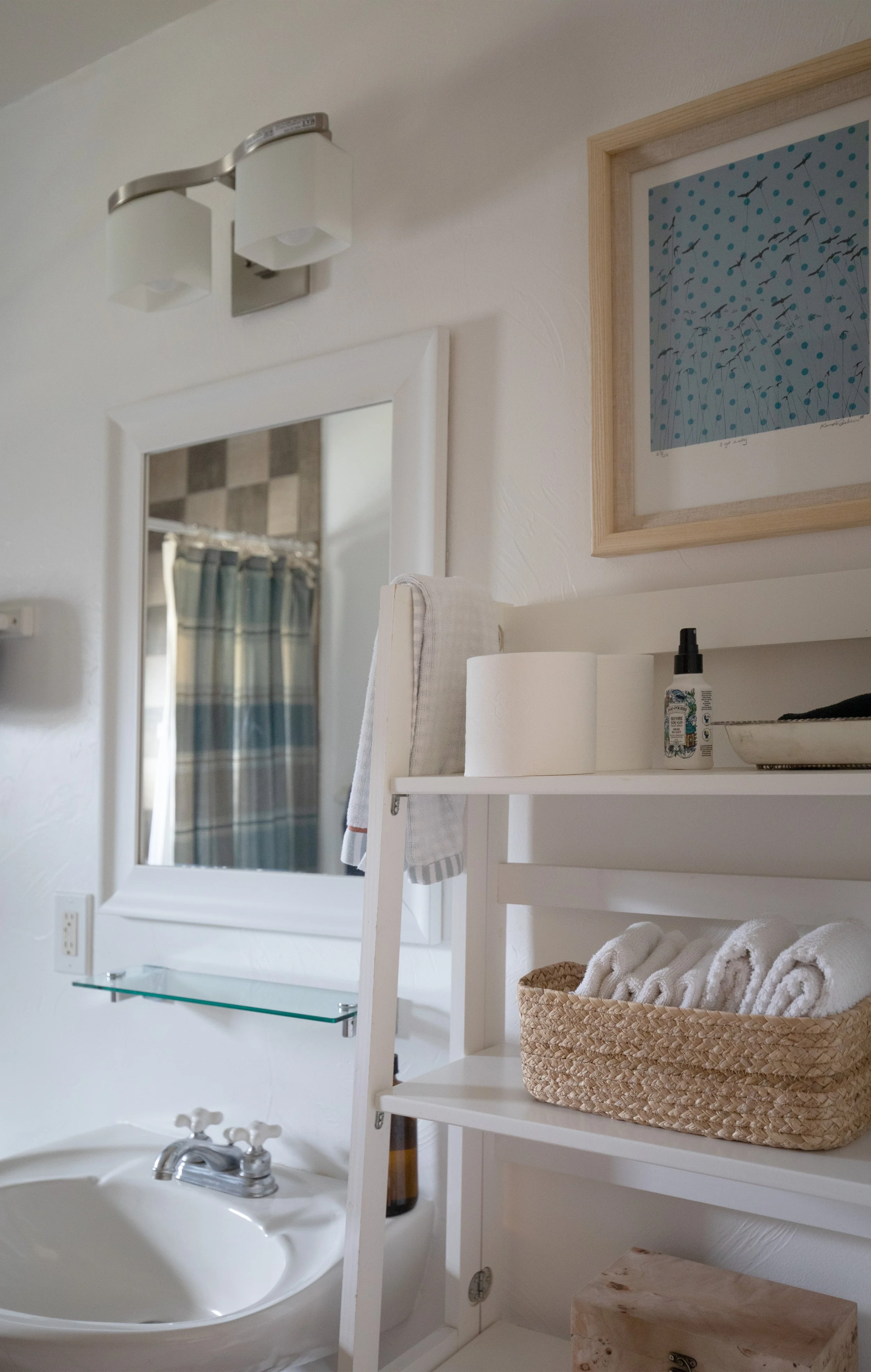 A bathroom with a white sink, a mirror, a towel hanging on a ladder, and a shelving unit with towels, toilet paper, and toiletries. There is a framed art piece on the wall and a ceiling light fixture. bozeman montana two dot interiors