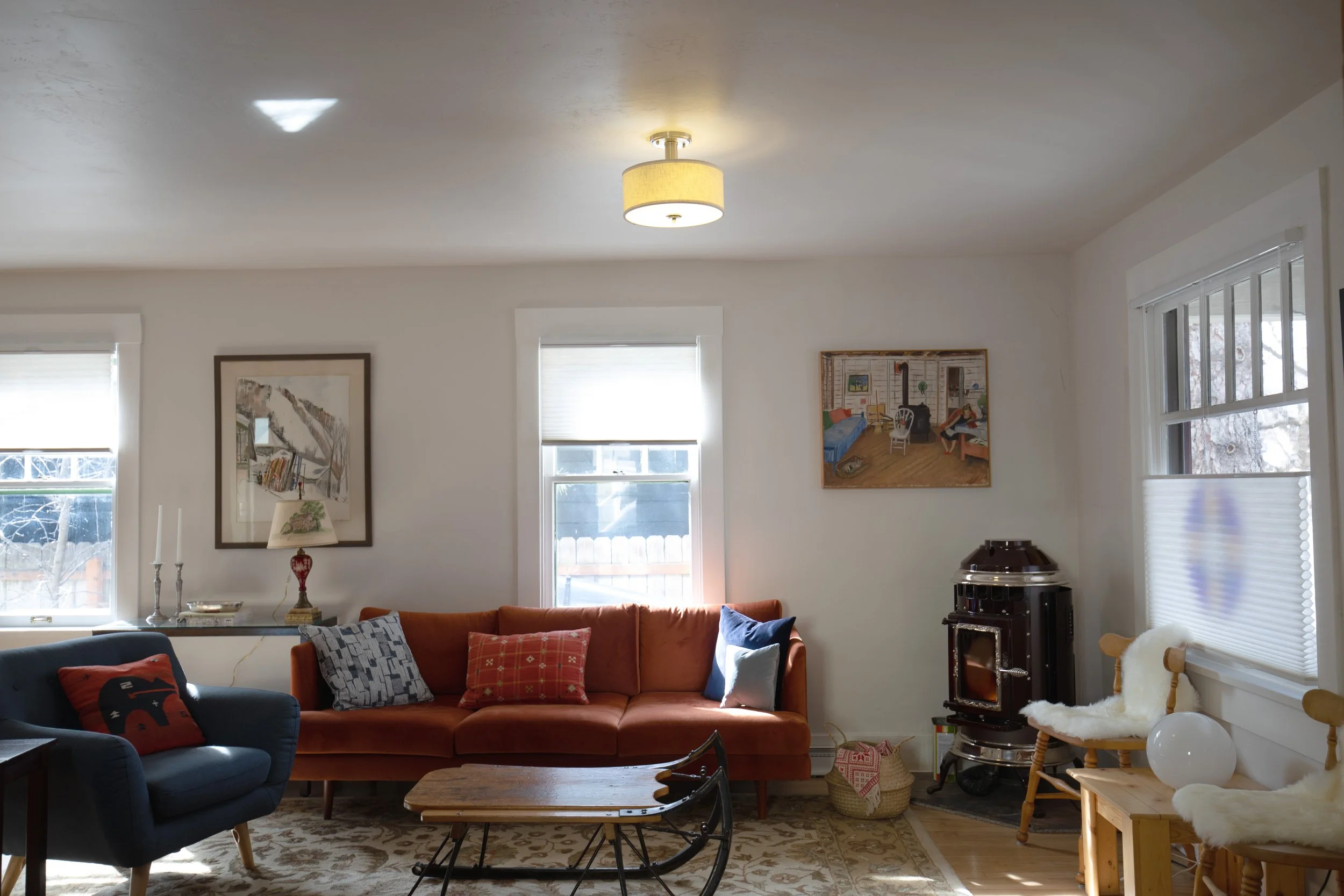 Living room with orange sofa, blue armchair, and wooden coffee table. Windows with blinds, wall art, stove, and cozy chairs with fur cushions. refresh renovation remodel interior designer bozeman montana two dot interiors