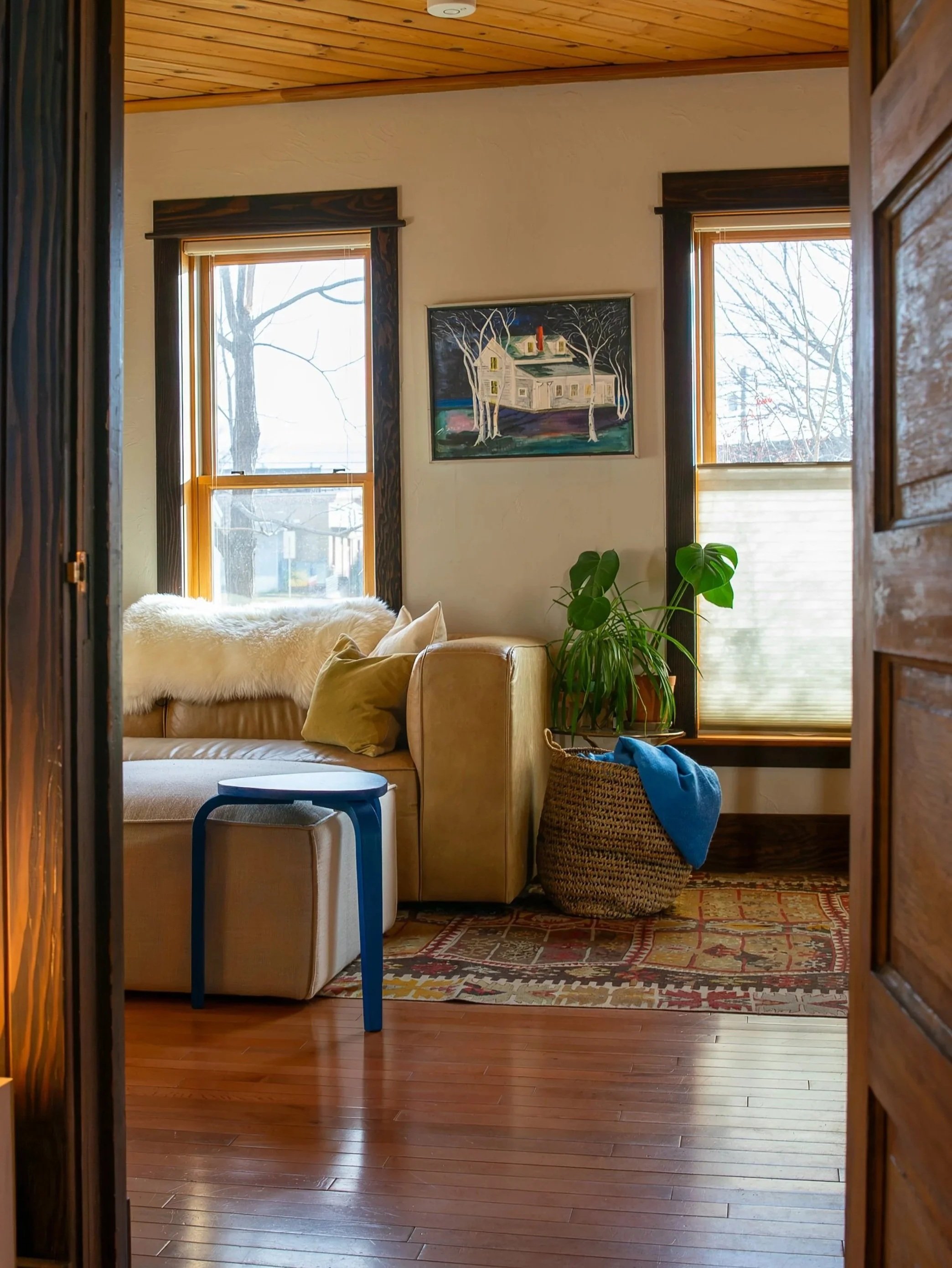 A cozy living room scene viewed from a doorway. It features a beige sofa with pillows, a textured rug on wooden floor, a small blue side table, and a woven basket with a blanket. Bozeman interior design, interior renovation, and interior styling. 