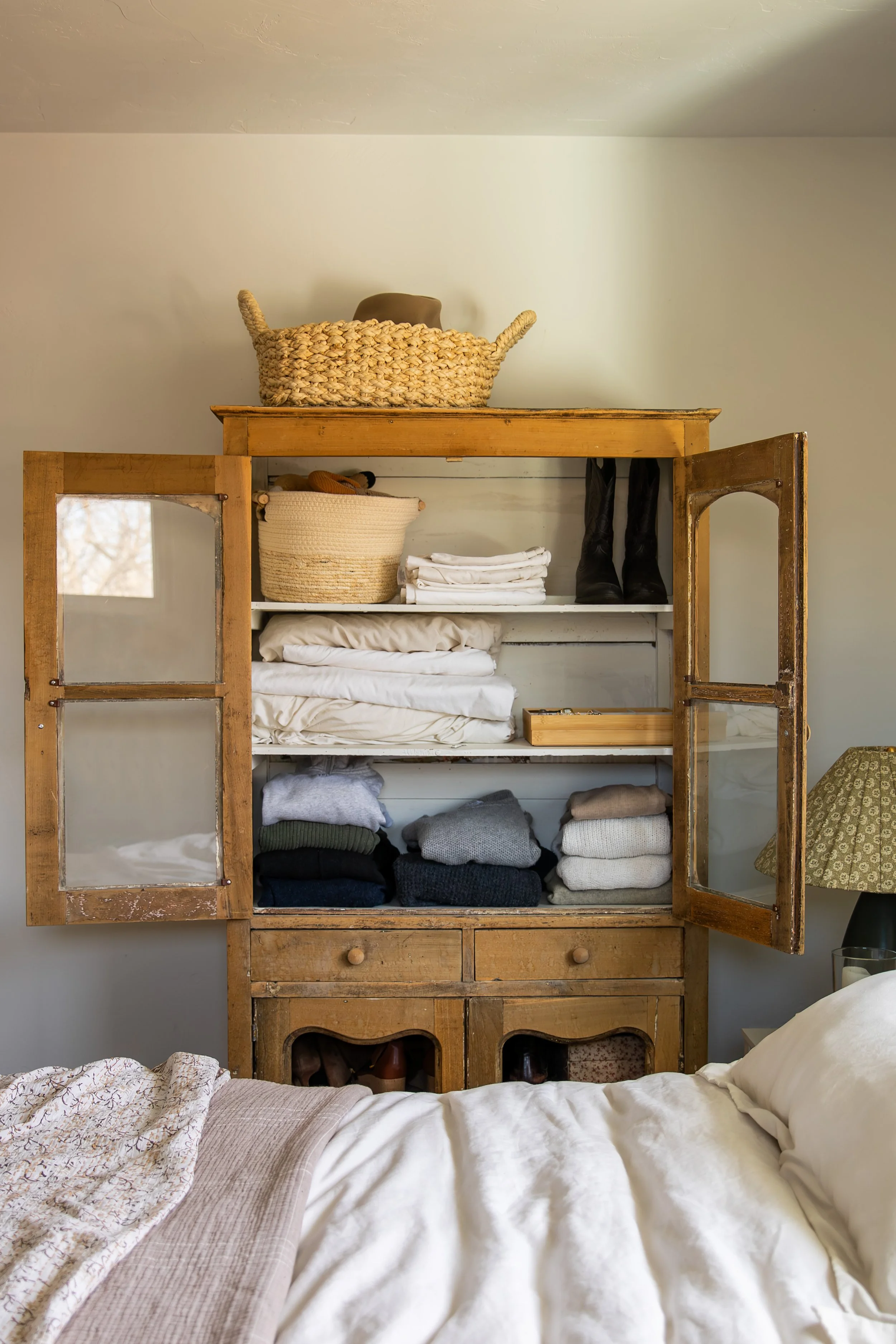 A wooden cabinet with glass doors filled with folded sweaters, a basket, and a pair of black boots. On top of the cabinet, there is a large woven basket. Bozeman interior design, interior renovation, and interior styling. 