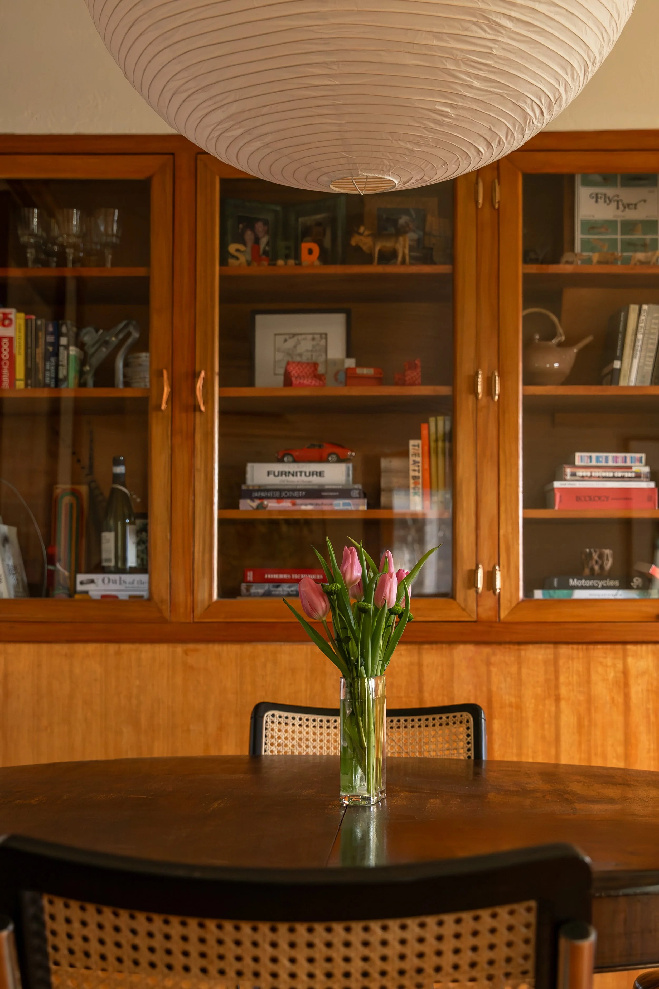A dining room with a dark wood table and a glass vase on top. In the background, there's a wooden cabinet with glass doors filled with books, decorative objects, and framed photos. Bozeman interior design, interior renovation, and interior styling. 
