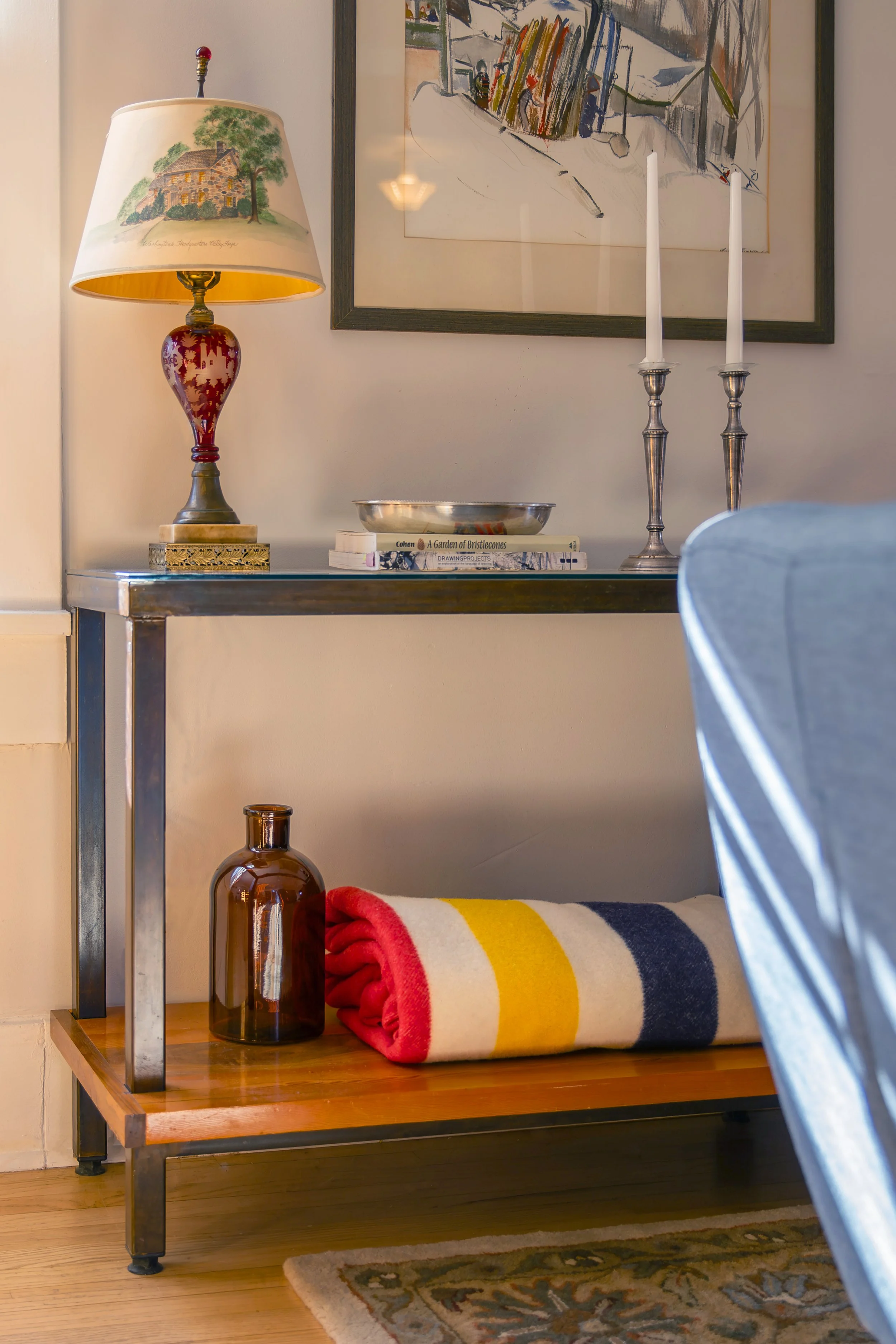 A decorative table with a glass top and metal frame, holding a colorful lamp with a landscape painting lampshade, a silver bowl, and a stack of books. A rolled red, yellow, white, and navy blanket is placed on the bottom shelf, next to a brown glass 