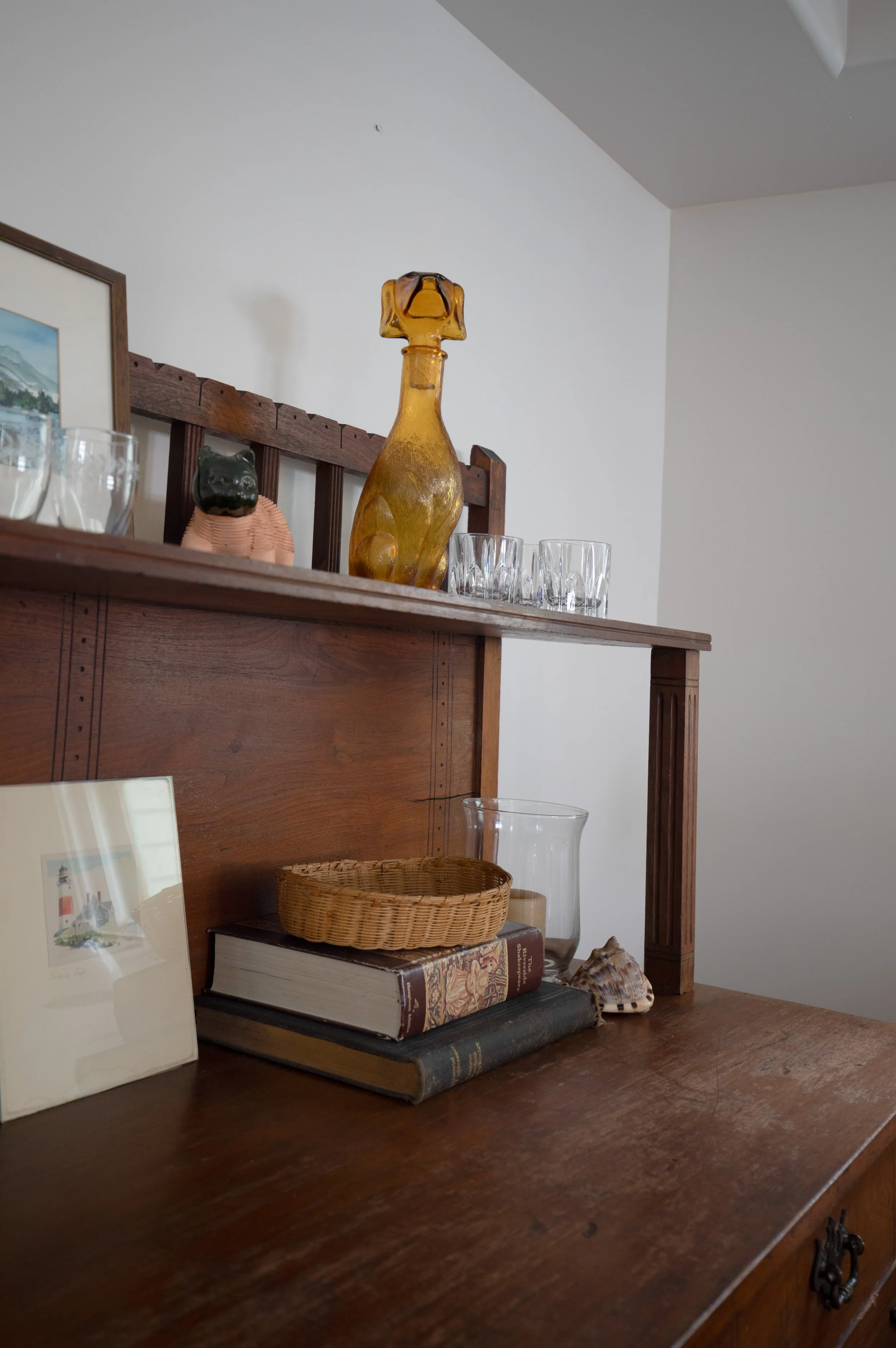 A wooden dresser with decorative items including a glass dog figure, a small bear figure, a framed picture, a wicker basket, two books, a glass vase, and a shell, with a white wall background. refresh renovation remodel interior designer bozeman mt