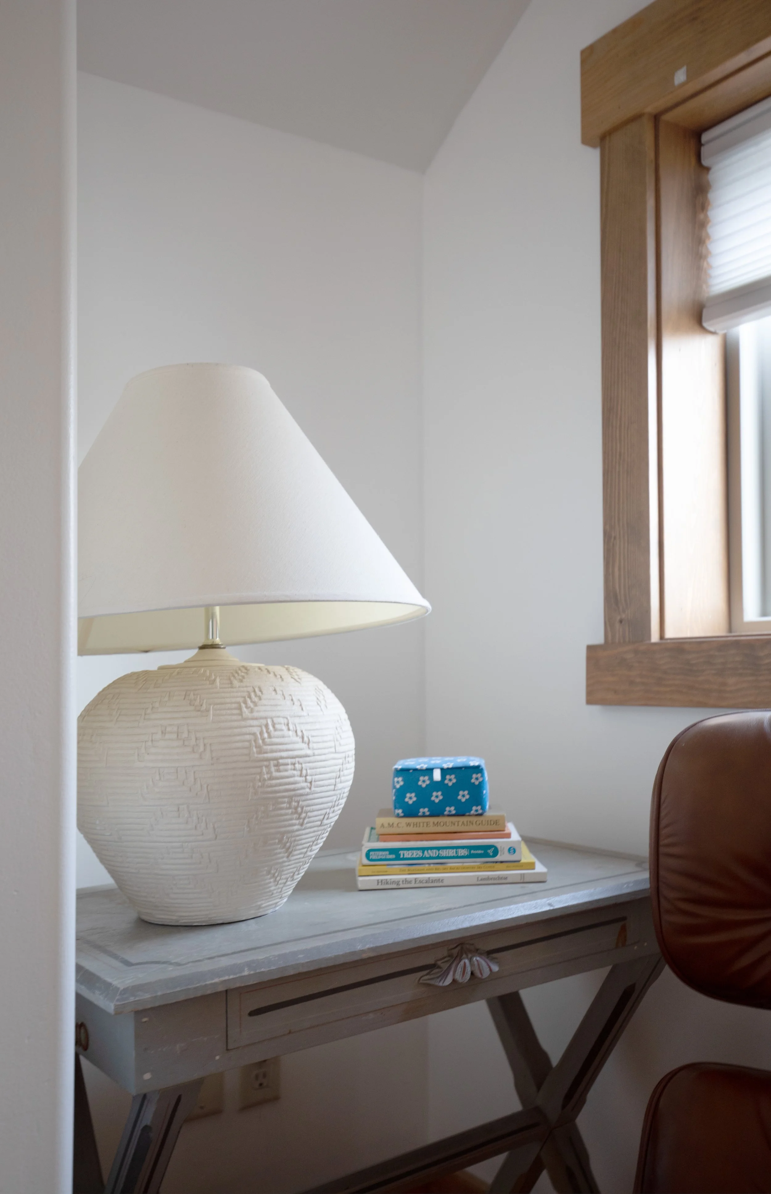 A desk with a large white textured lamp, a small blue box, and several books, in front of a window with wooden trim. refresh renovation remodel interior designer bozeman montana two dot interiors