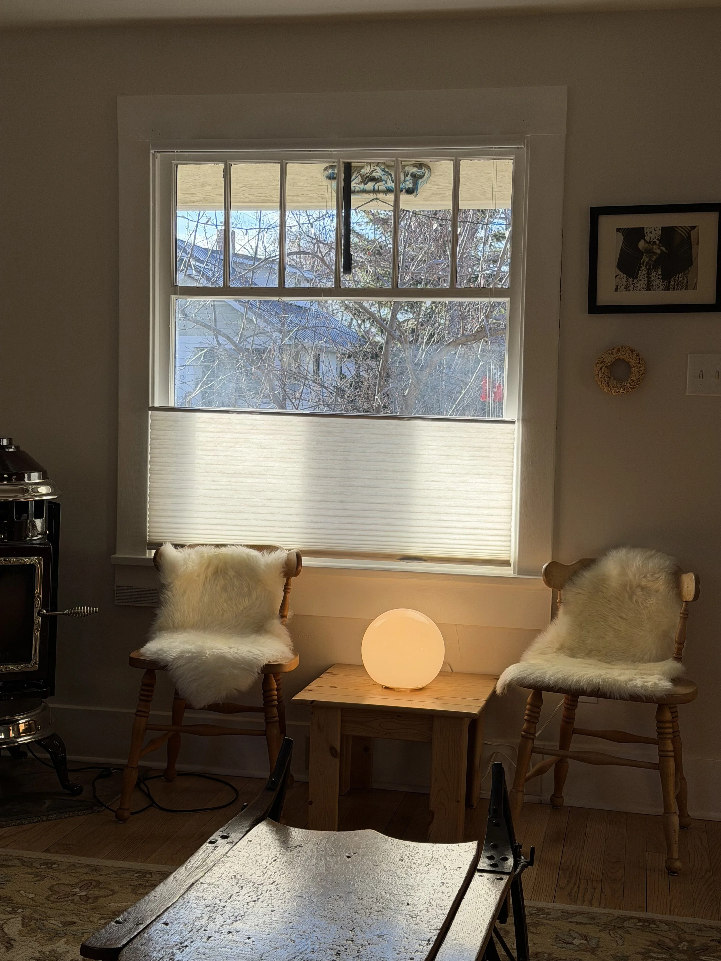 A cozy interior scene with two wooden chairs draped in white sheepskin, a small wooden table with a glowing orb lamp between them, and a window with blinds partially open showing trees outside.  Bozeman Montana Interior Design Affordable ikea 