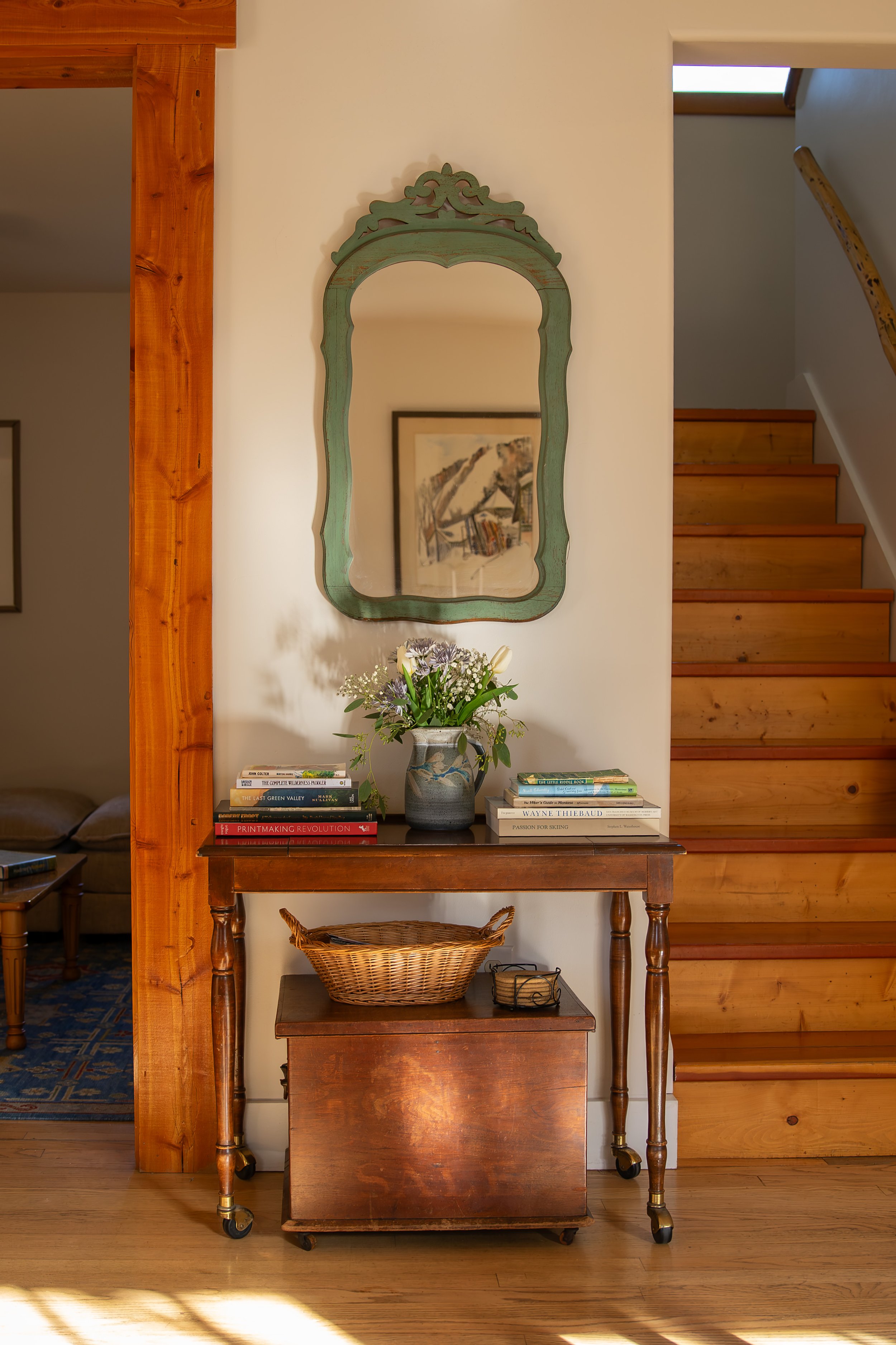 A wooden console table with a vase of flowers, books, and a wicker basket in front of a white wall with a decorative mirror. A staircase with wooden steps is on the right side.