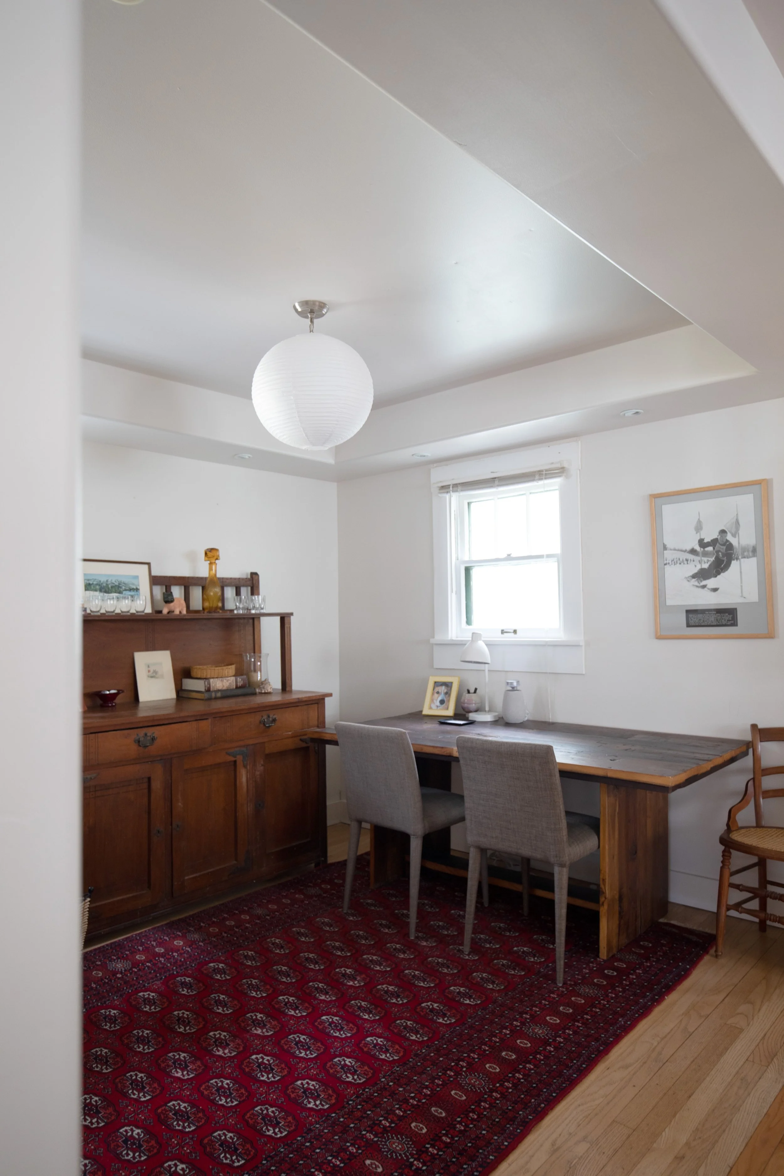 Interior of a dining room with a wooden table, gray chairs, a wooden sideboard, a framed photo, a white window, a framed black-and-white sports photo, and a red patterned rug. STR refresh interior design bozeman montana two dot interiors