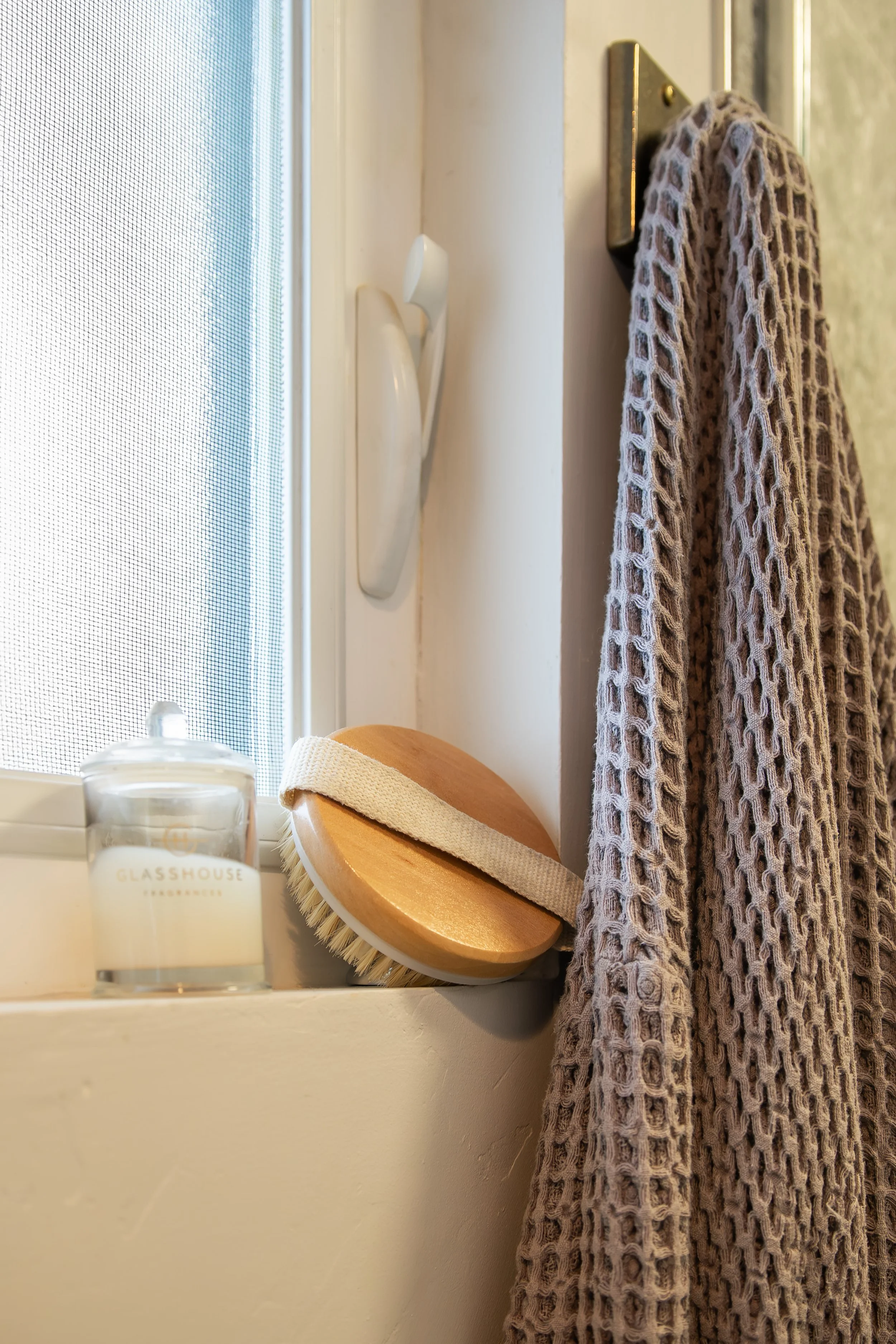 A bathroom window sill with a candle, a body brush, a hook with a knit towel hanging on it, and a window. Bozeman interior design, interior renovation, and interior styling. 