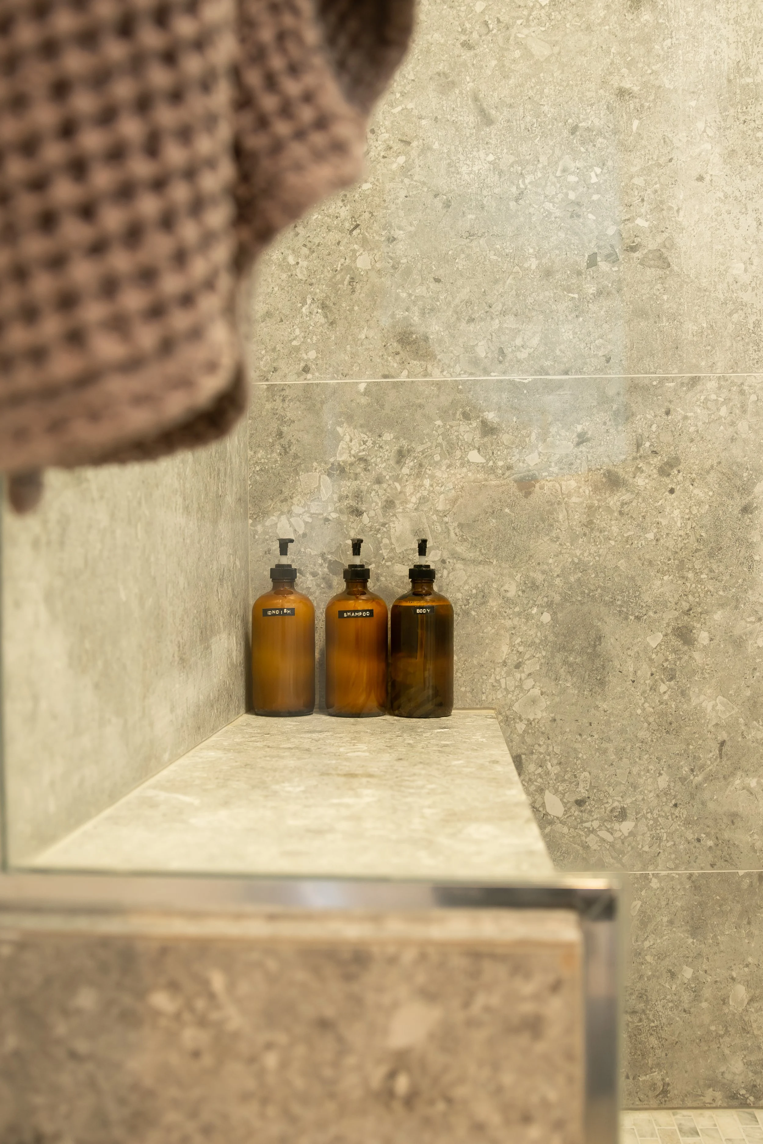 Three brown amber bottles labeled 'shampoo,' 'body,' and 'conditioner' sit on a beige marble ledge in a shower with gray textured tile walls. Bozeman interior design, interior renovation, and interior styling. 