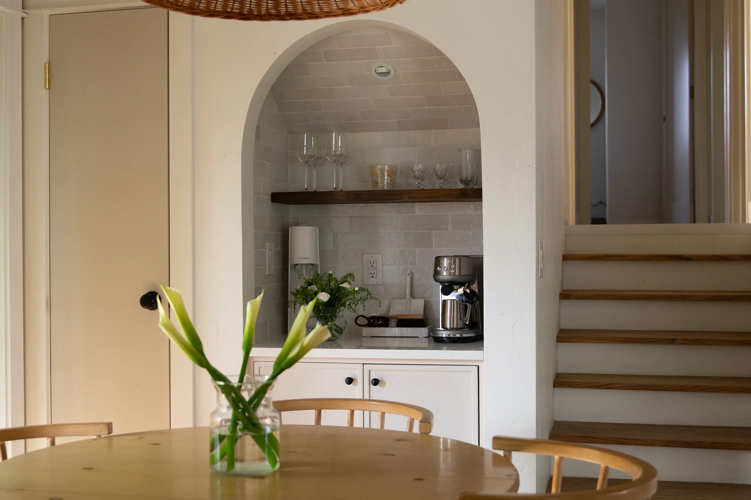 Interior view of a kitchen with a small round dining table, a vase with lilies, and an open archway revealing a coffee station with a coffee maker, glassware on a shelf, and a plant. Bozeman interior design, interior renovation, and interior styling.