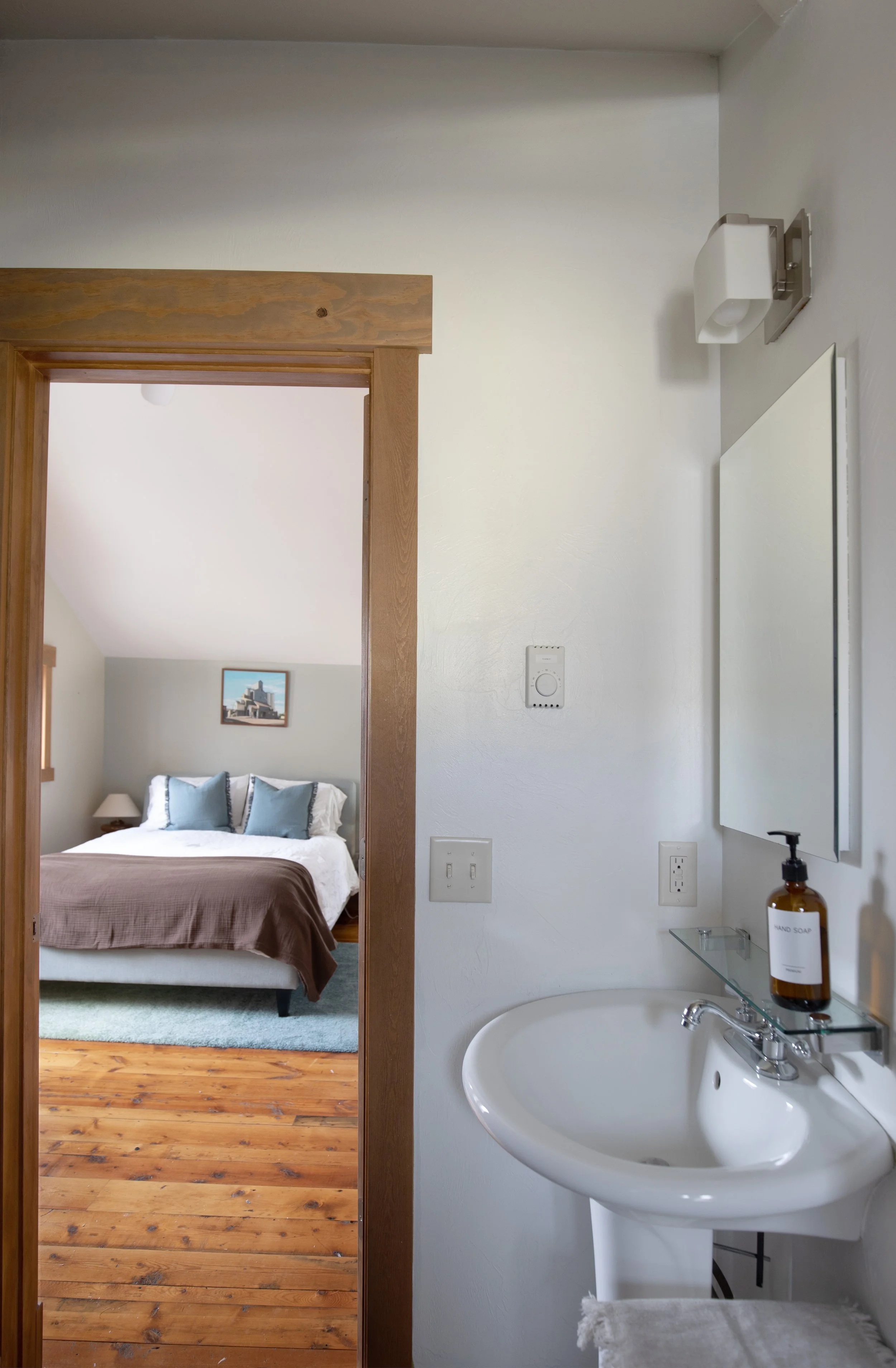 View of a bedroom through a doorway, with a bed and pillows, and part of a bathroom with a sink, mirror, soap dispenser, and light fixture. STR refresh service interior design bozeman montana two dot interiors