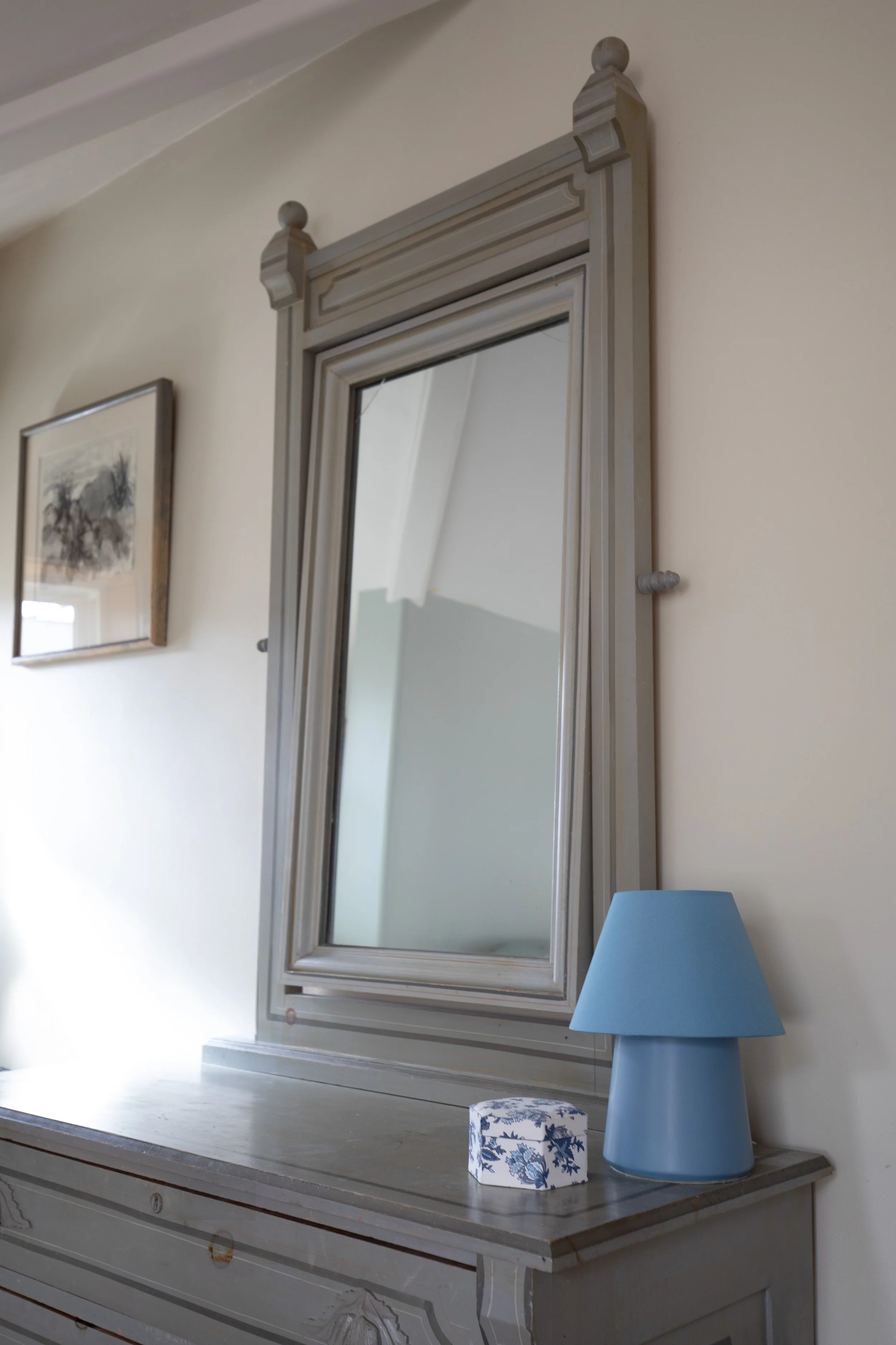 A decorative wooden dresser with a large mirror, a blue lamp, and a small patterned box, against a light-colored wall with framed artwork. bozeman montana two dot interiors