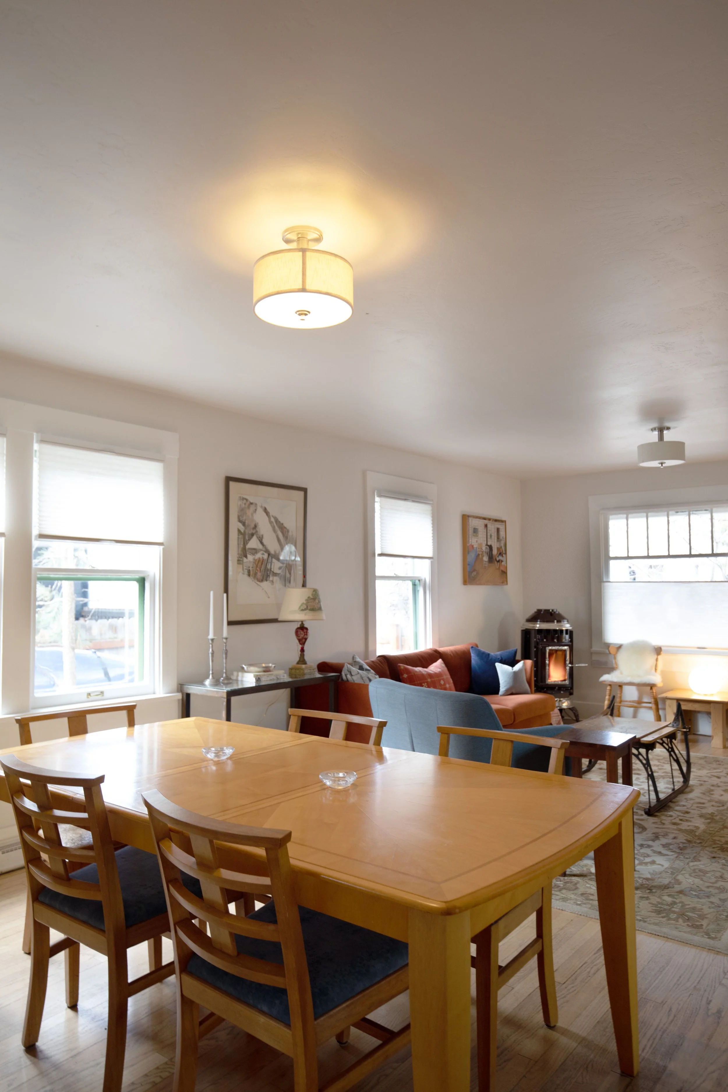 A living room with a wooden dining table and six chairs, a sofa, a coffee table, a wood stove, and multiple windows with blinds.