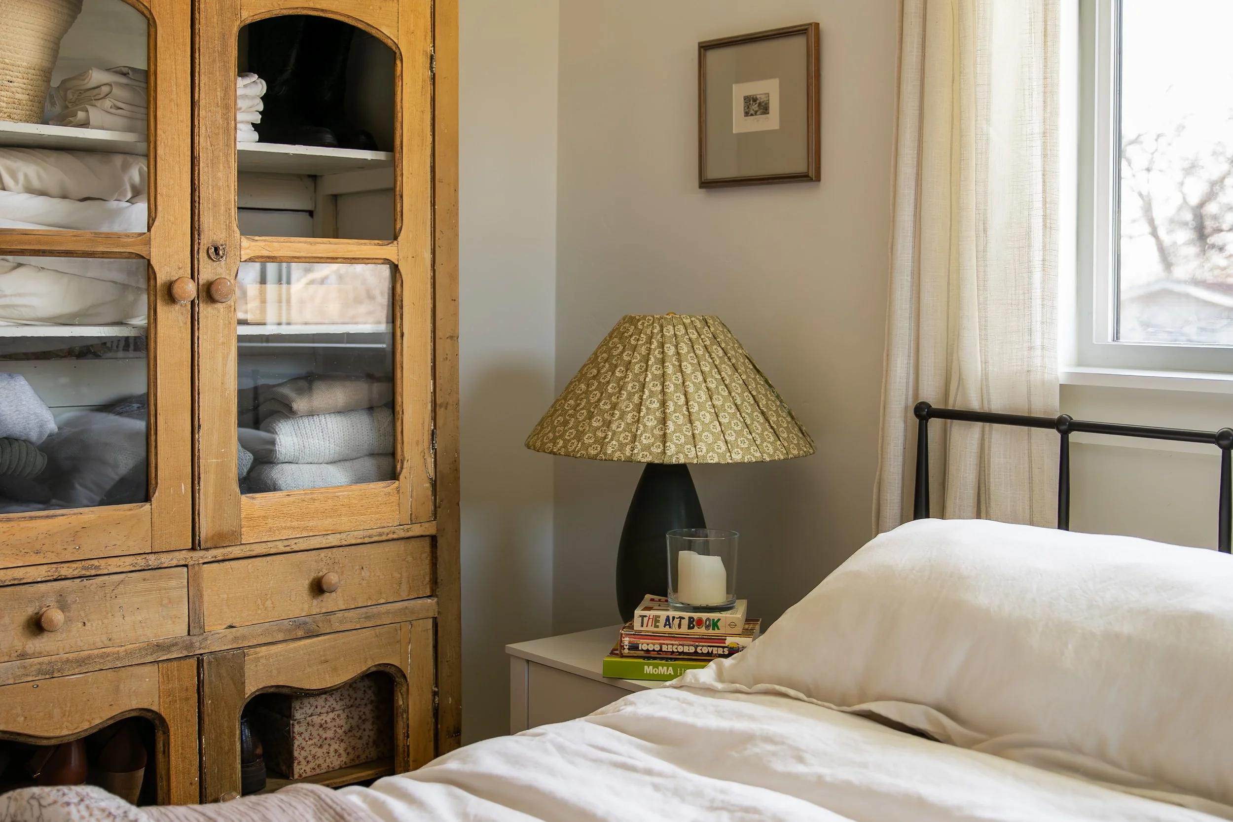 A cozy bedroom corner with a wooden cabinet filled with neatly folded laundry, a side table, and a window with light curtains letting in natural light. Bozeman interior design, interior renovation, and interior styling.