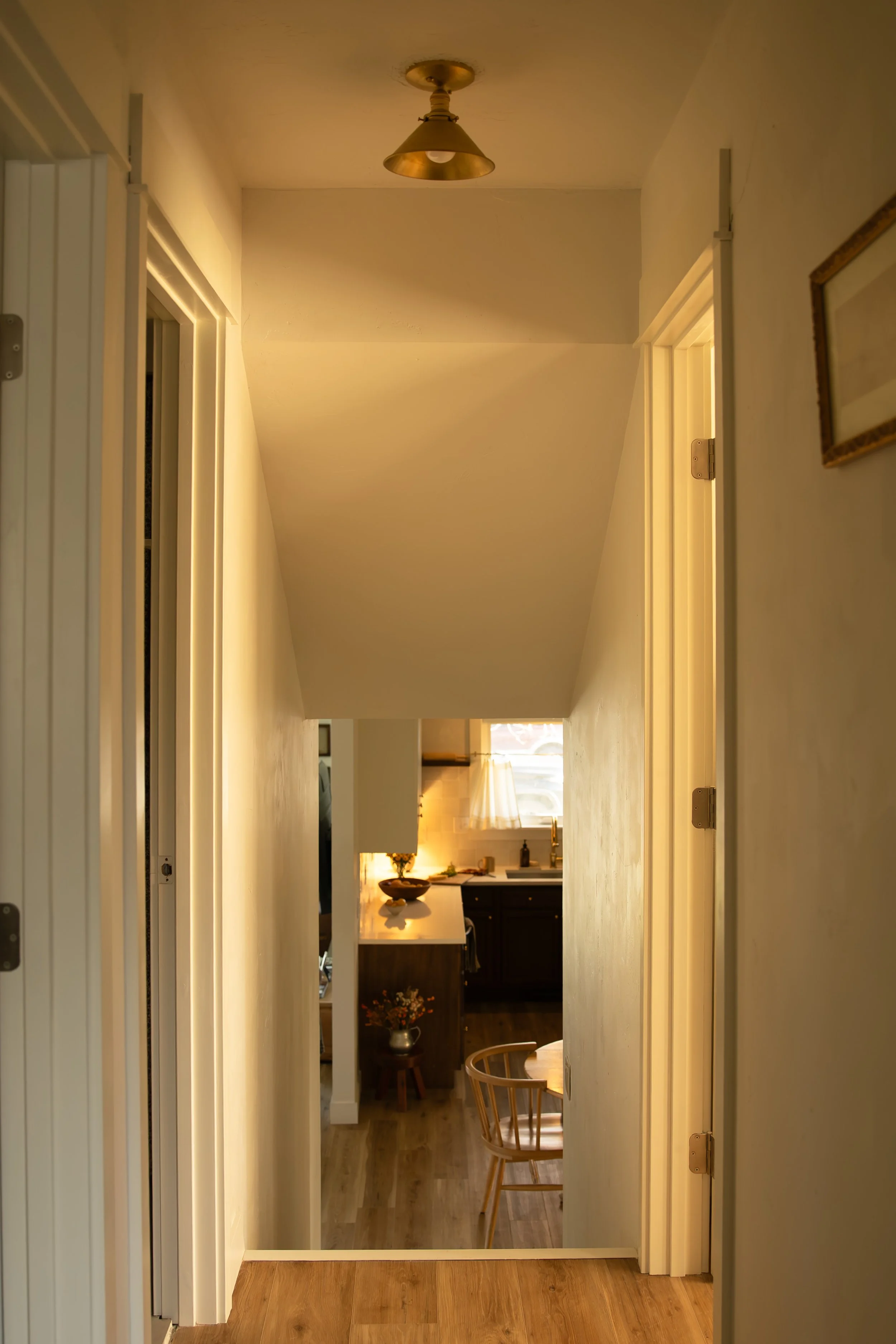 A view of a narrow corridor leading into a kitchen area with wooden flooring, a dining table with wooden chairs, and kitchen cabinets with a window above the sink. Bozeman interior design, interior renovation, and interior styling.